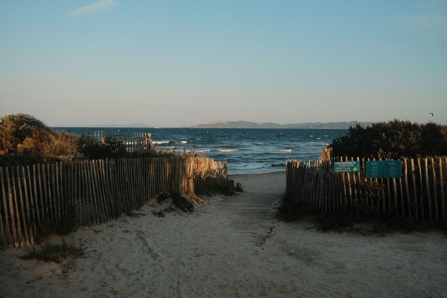 Sandy beach path with wooden fences leads towards the ocean under a bright blue sky.