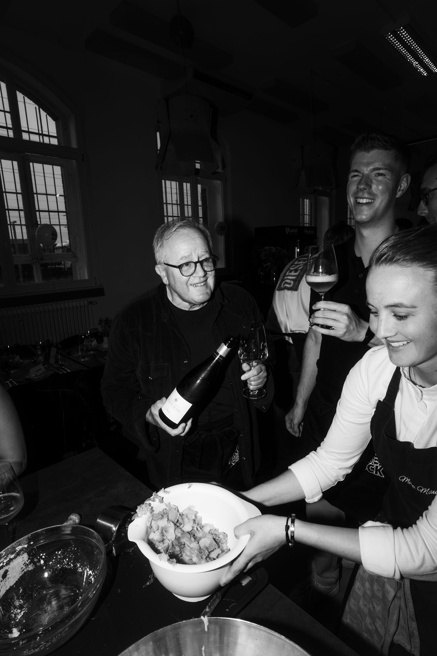 Black and white photo of people gathering around food and drinks in casual indoor setting.