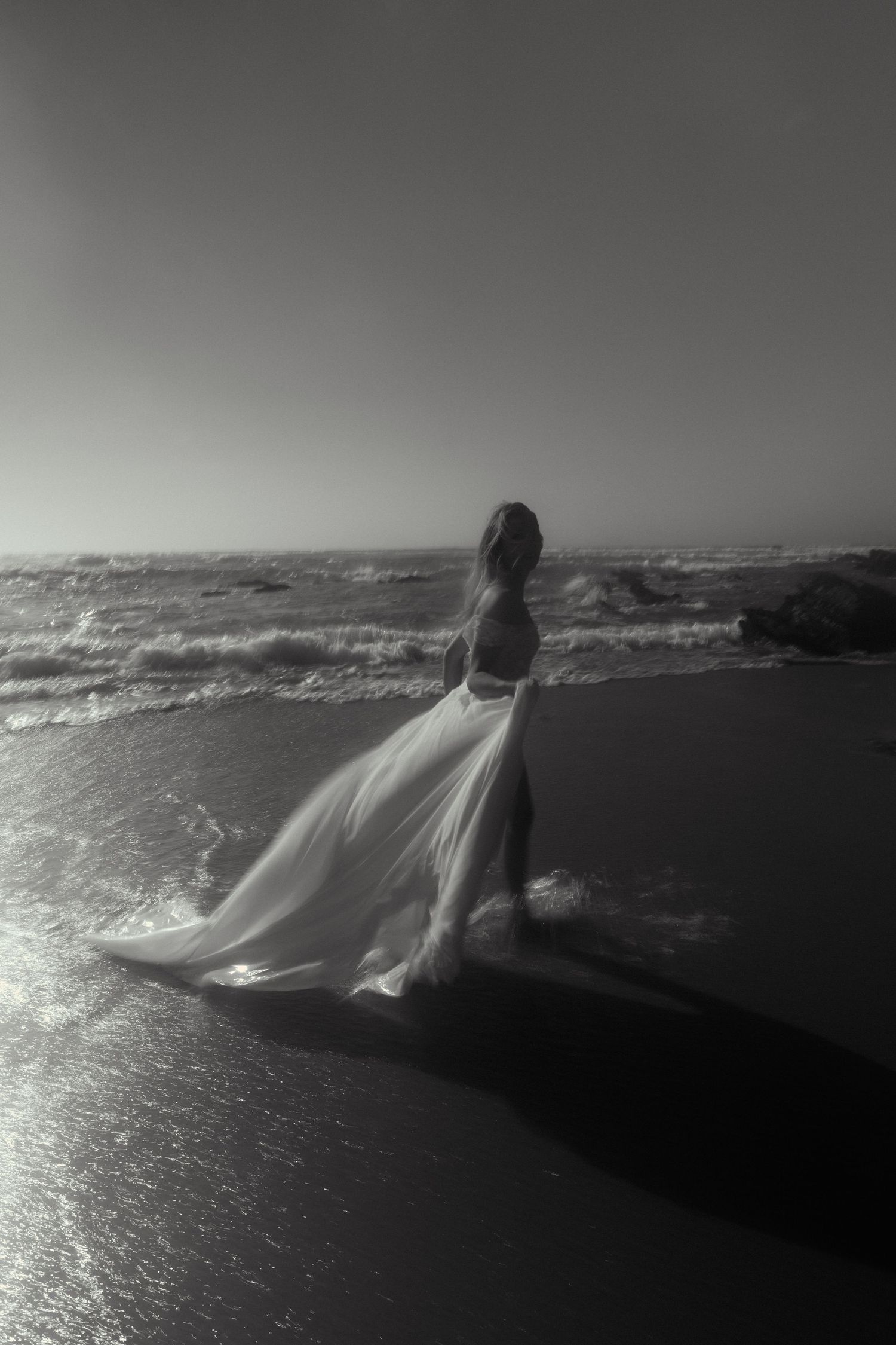 Dramatic beach scene with flowing white wedding dress catching sunlight against dark waters.
