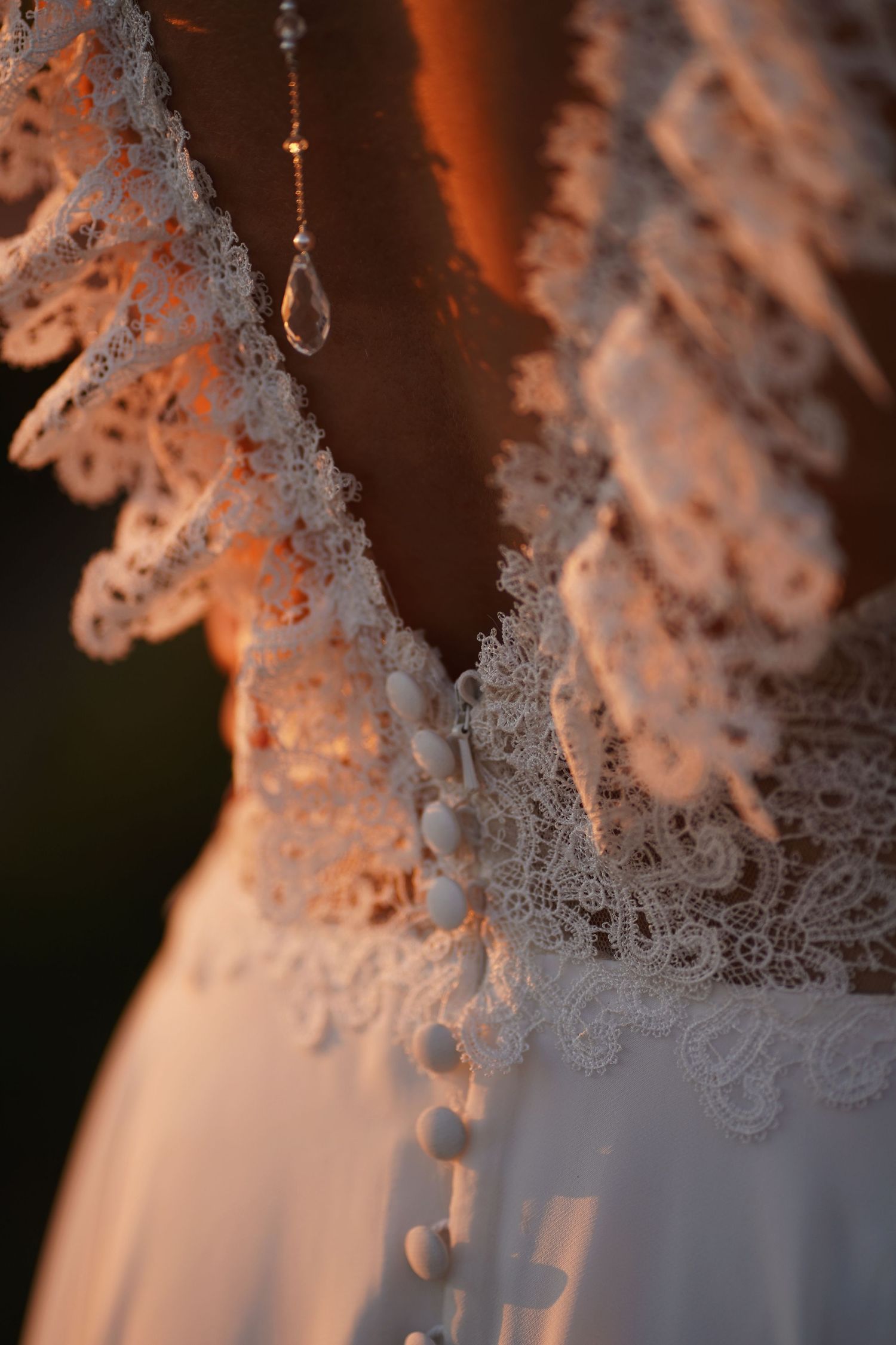 Intricate lace detail and crystal pendant on a wedding dress catching warm light.