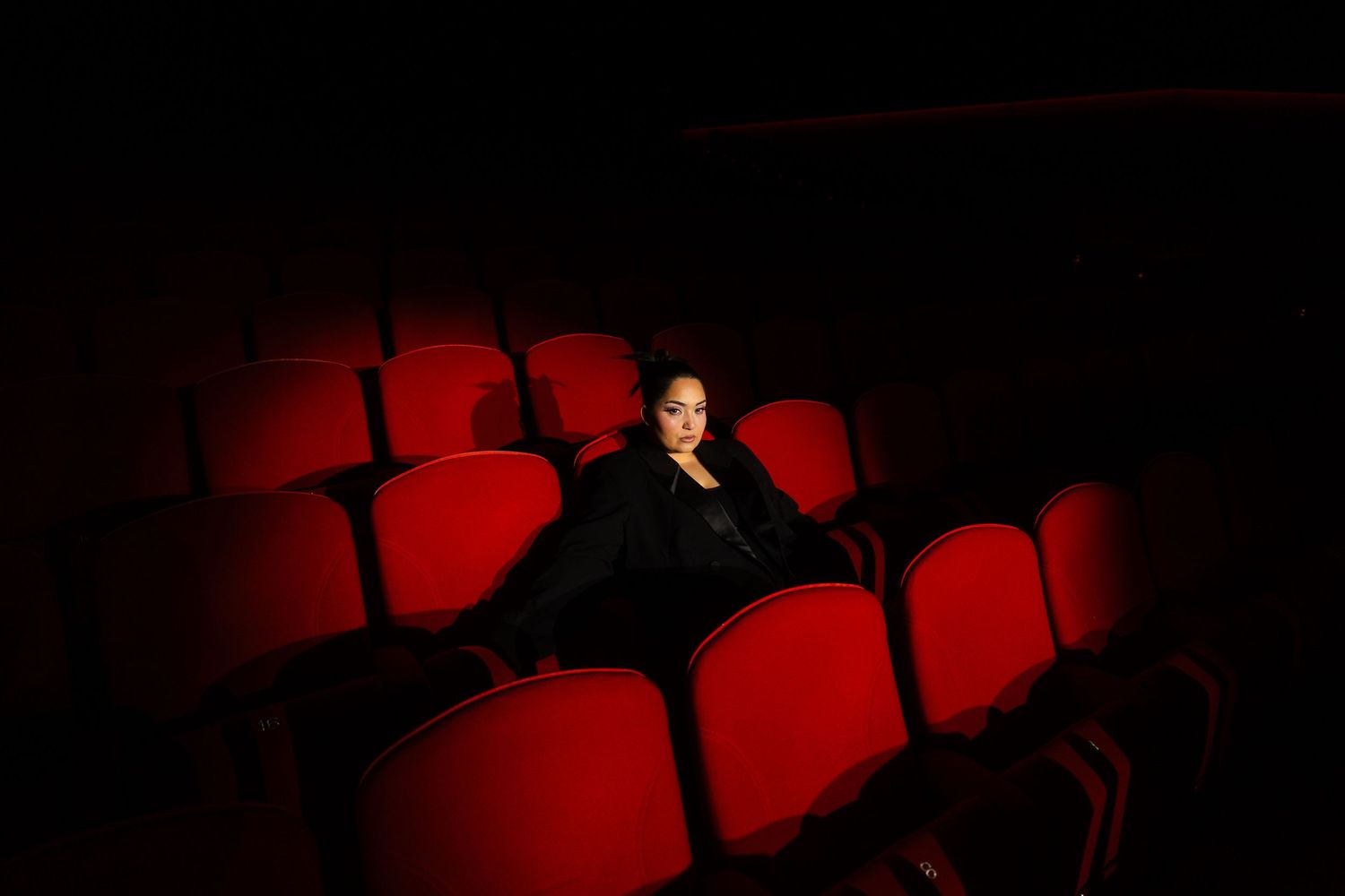 Bahar Kizil seated alone in empty theater with dramatic red lighting on vintage cinema seats, atmospheric cinema ambiance.