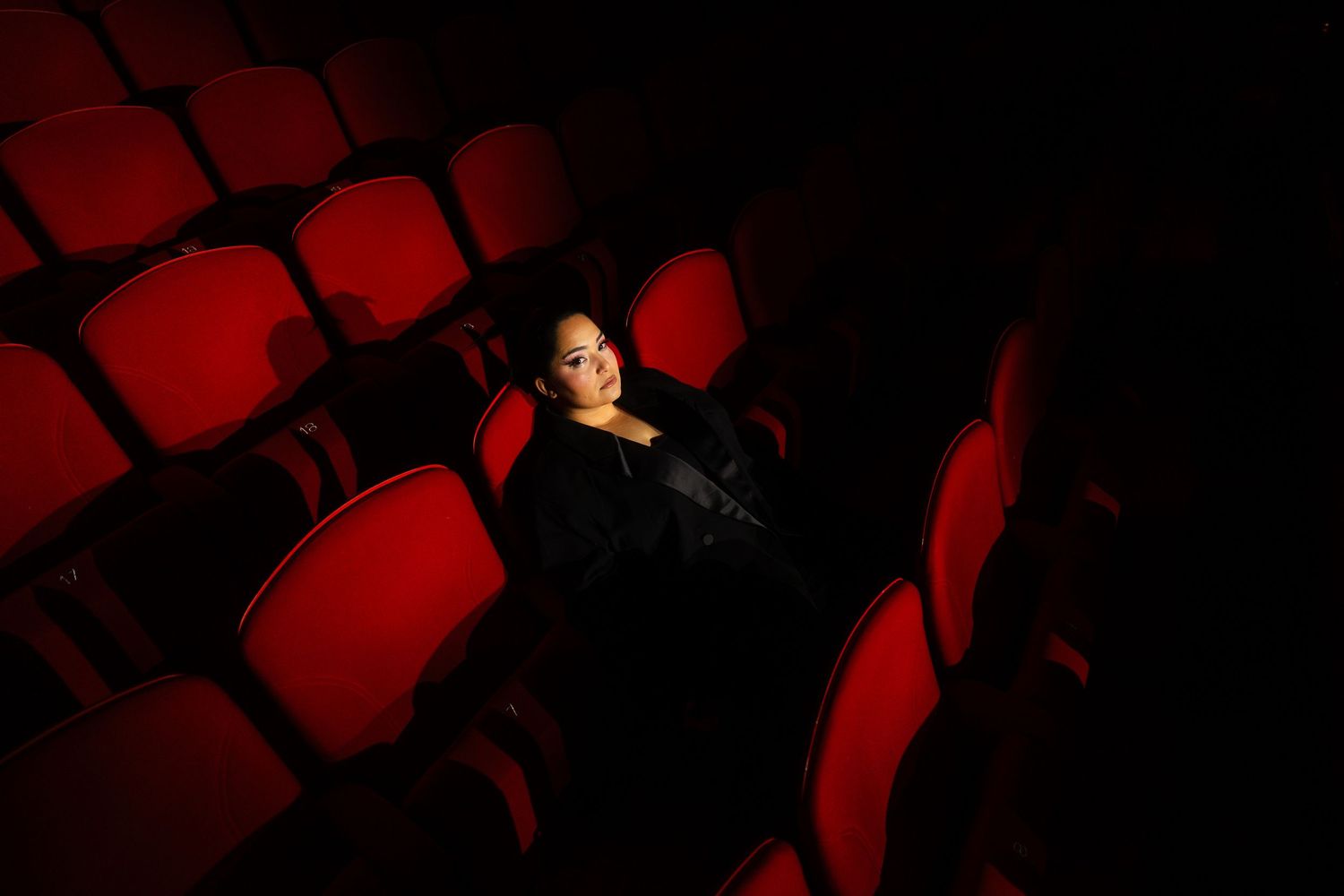 Bahar Kizil seated alone in empty theater with dramatic red lighting on vintage cinema seats, atmospheric cinema ambiance.
