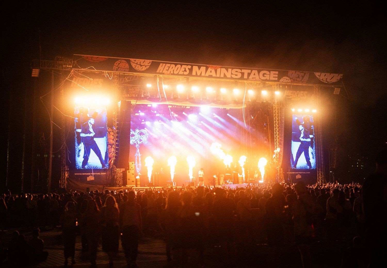 Concert stage lit up with dramatic orange lighting and pyrotechnics creating a spectacular nighttime performance atmosphere.
