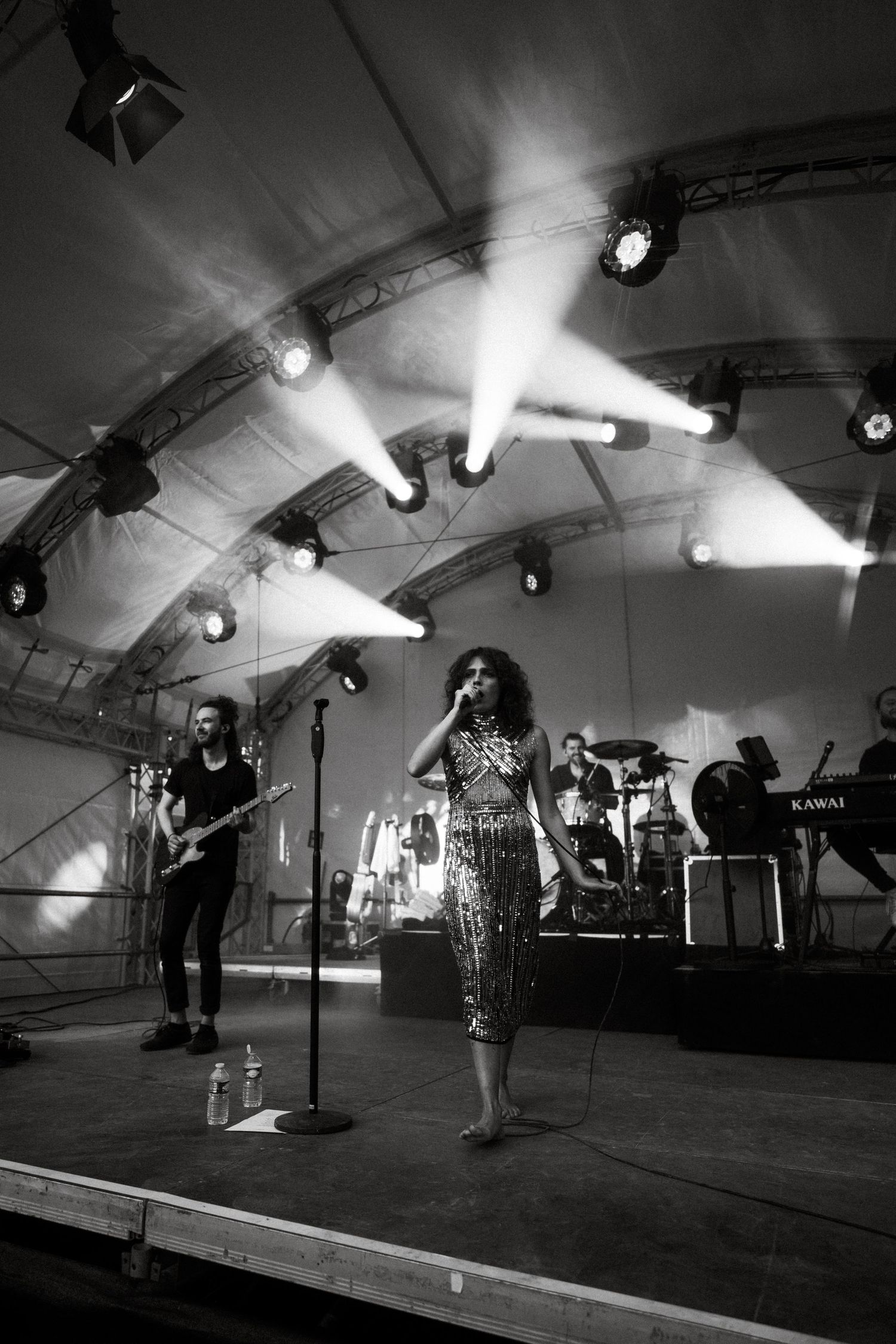 Concert performers on stage in a large tent with dramatic overhead lighting and equipment.