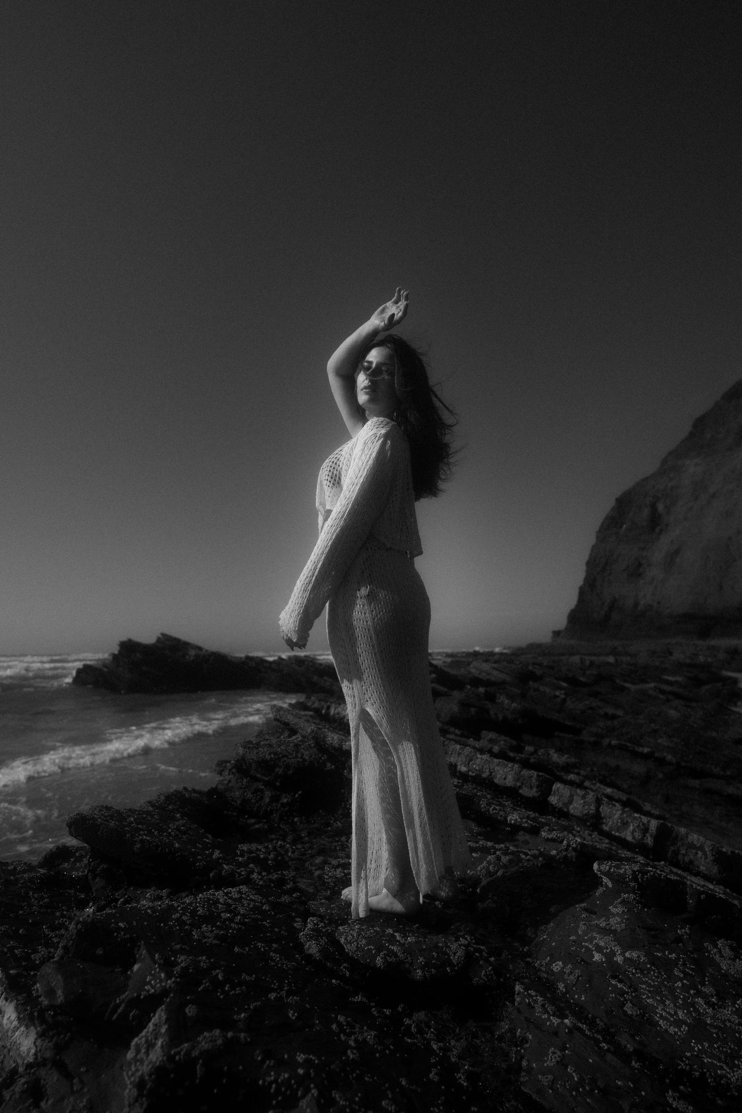 Ethereal figure in flowing white dress poses dramatically against coastal cliffs in black and white.
