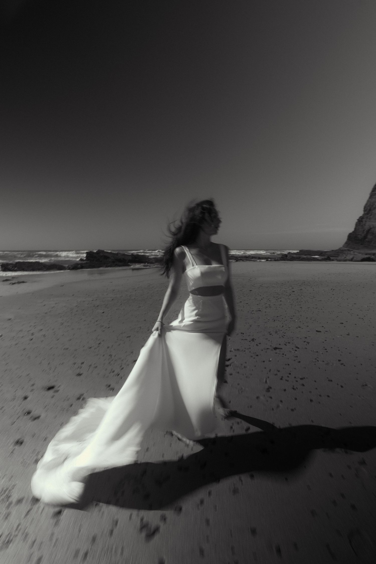 White wedding dress flowing in the wind on dramatic black and white beach scene.