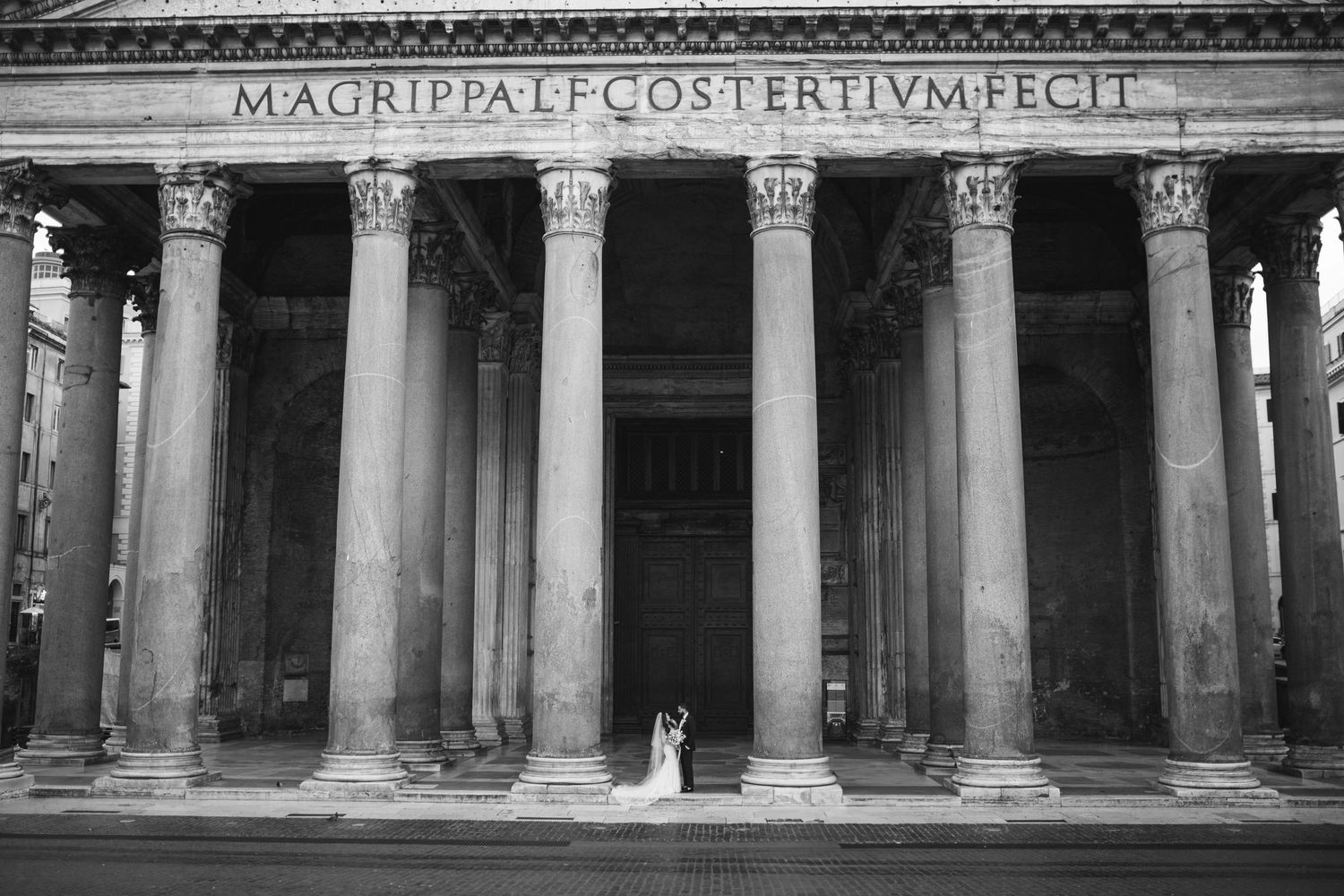 Black and white exterior photograph of a couple at the historic Pantheon with its grand Corinthian columns in Rome, Italy.