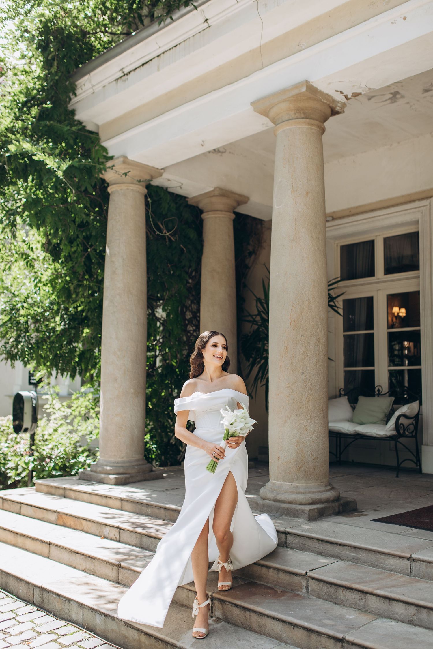 Elegant off-shoulder white wedding dress with thigh slit photographed on grand stone column porch with greenery.