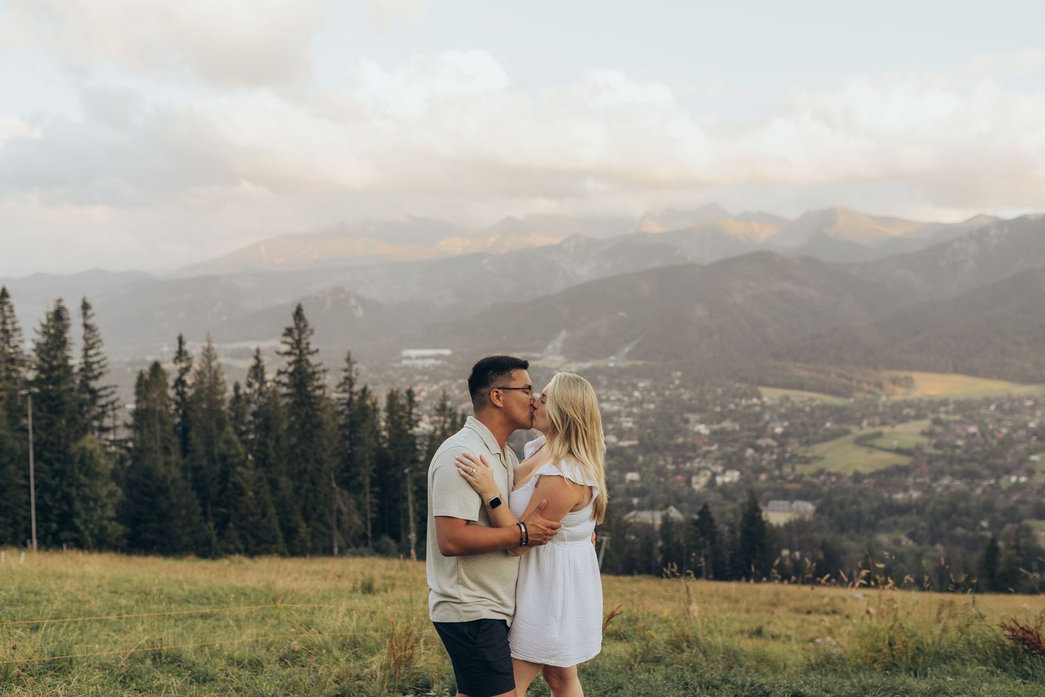 A couple shares a romantic moment on a hilltop meadow overlooking a mountain valley with pine trees at sunset.