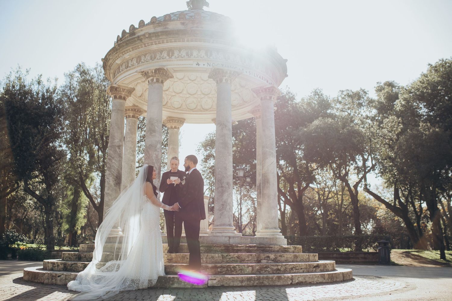 A wedding ceremony takes place under an elegant stone gazebo with sunlight streaming through the columns.