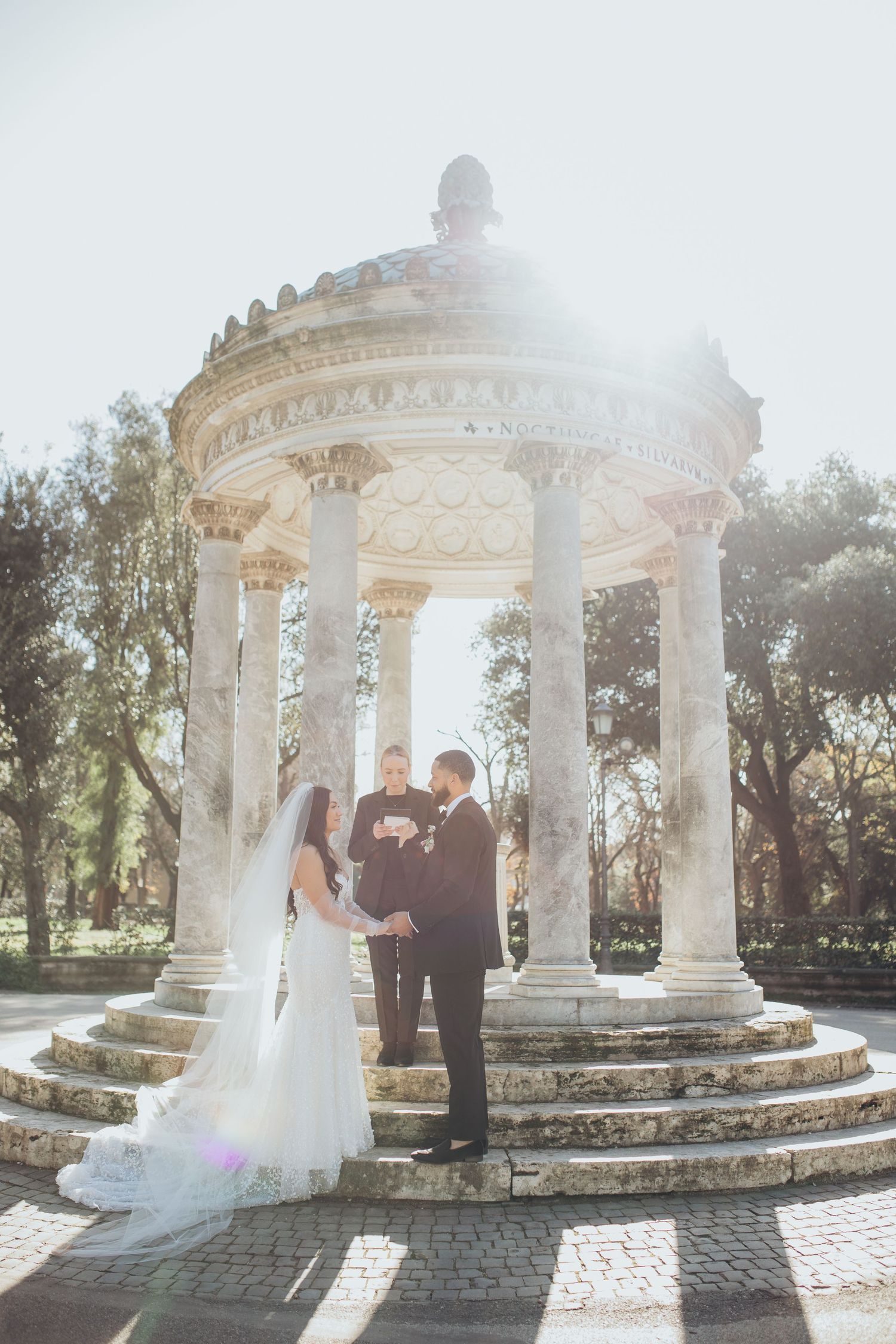 Elegant wedding couple embraces under ornate white marble gazebo with classical columns and sunlight streaming through.