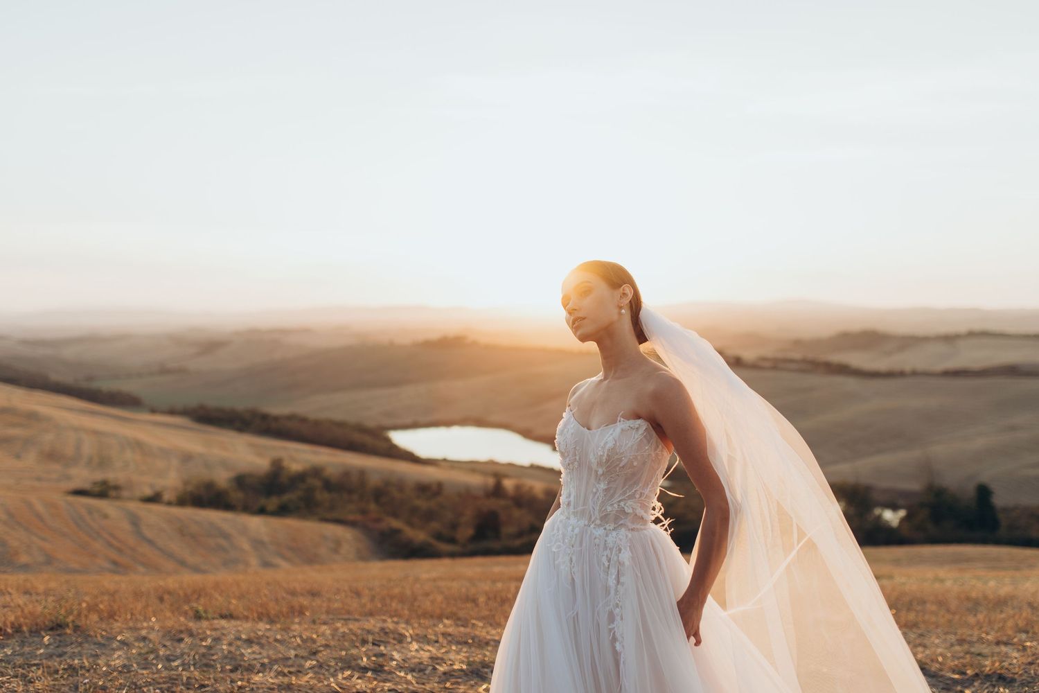 A bride in a flowing white dress and veil stands against rolling hills at golden hour sunset.
