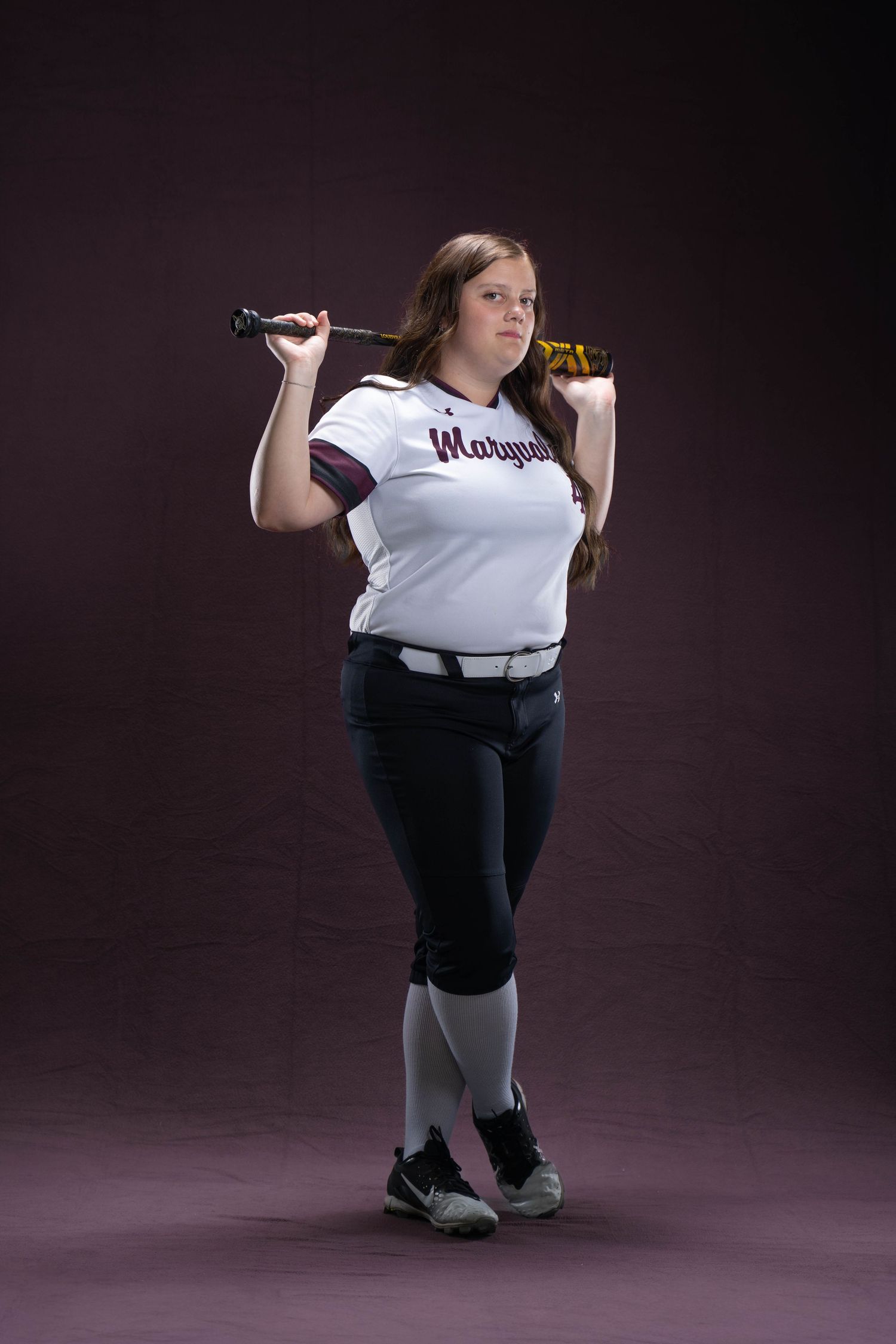 Young softball player in Marquette jersey poses with bat over shoulder against dark studio background.