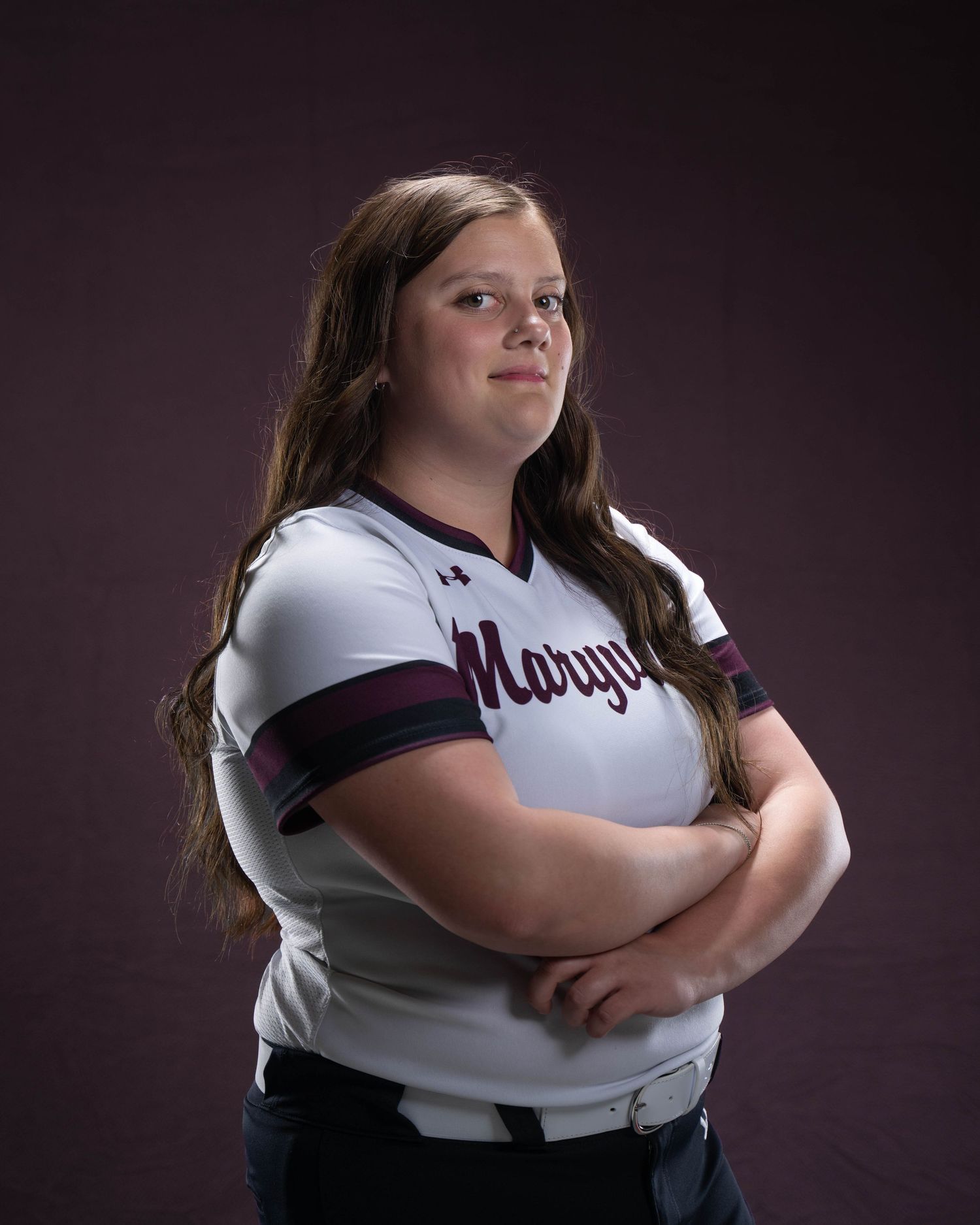 Young female softball player in white Marygold uniform poses with arms crossed against dark purple background.