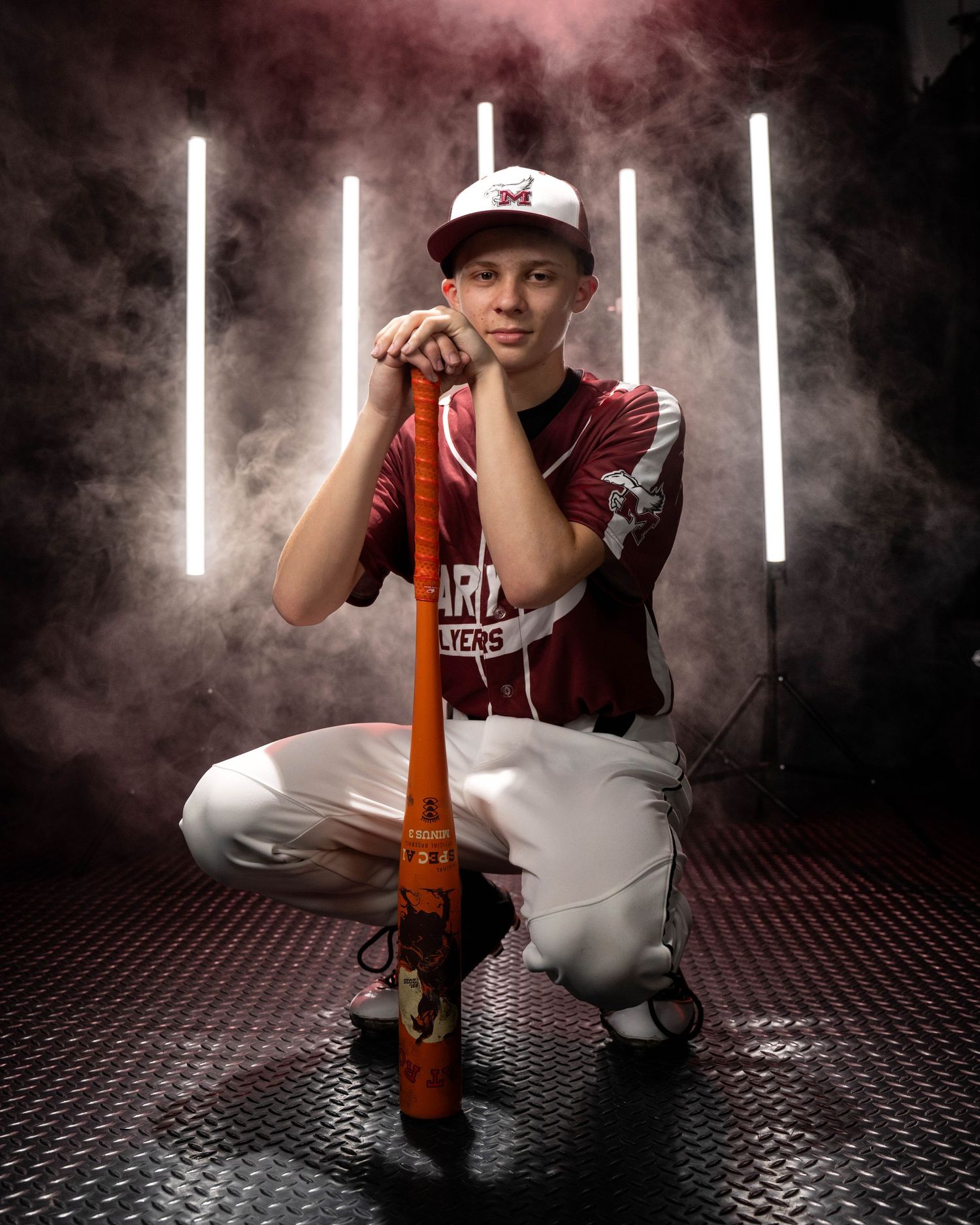A young baseball player in maroon uniform kneels with a bat in a dramatic smoky studio with neon lights.