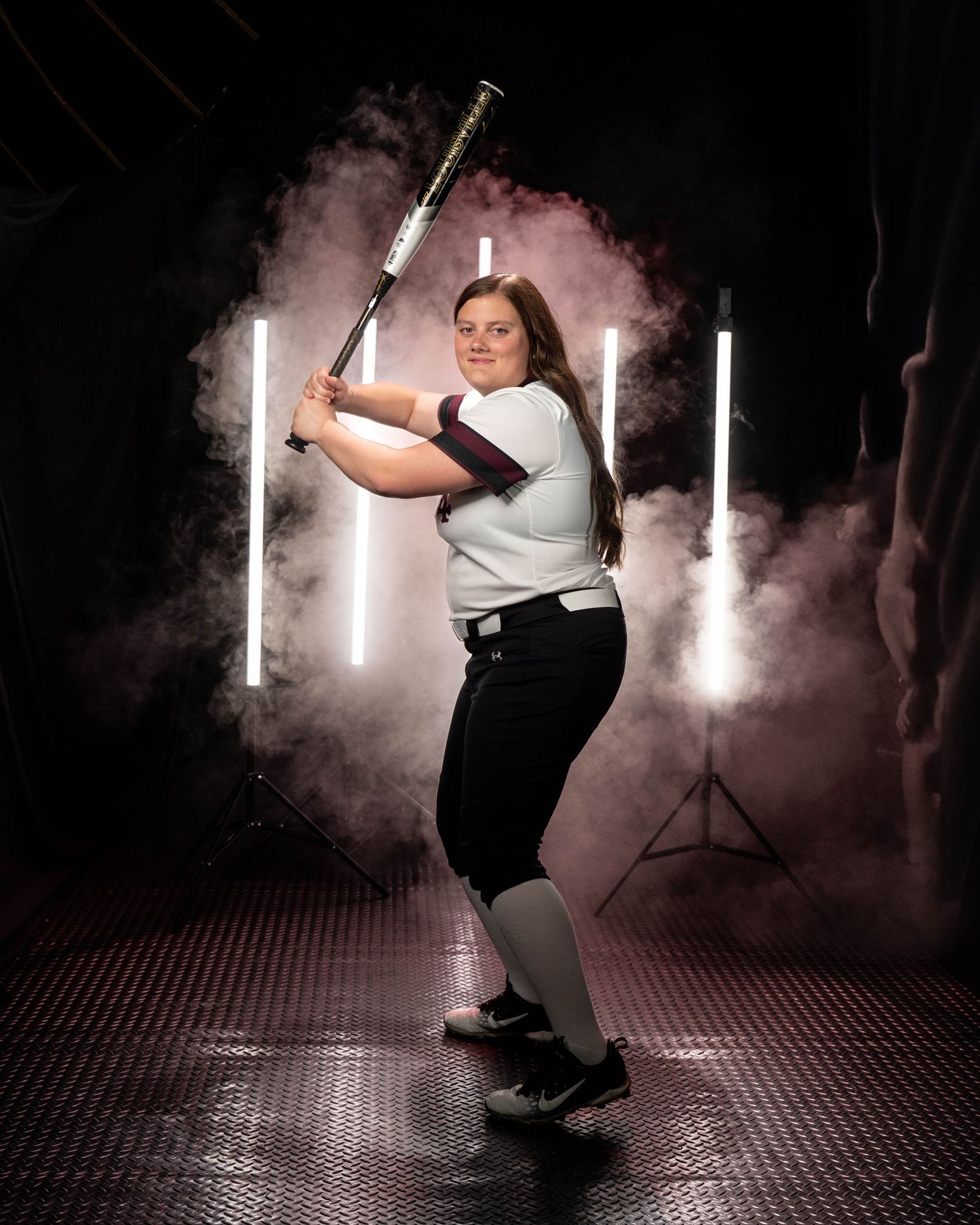 A female softball player in uniform stands in batting stance with a bat in a smoky studio with neon lights.