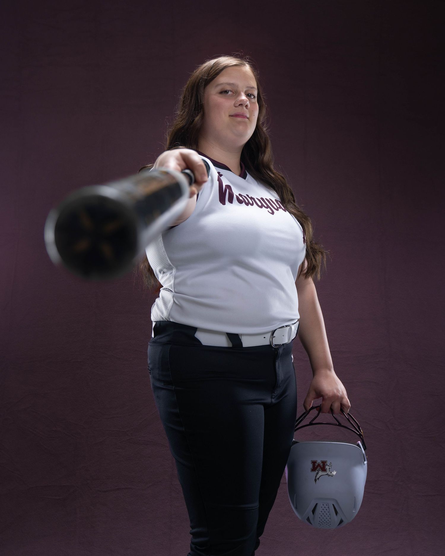 Young female softball player in uniform pointing bat at camera while holding helmet in studio portrait.
