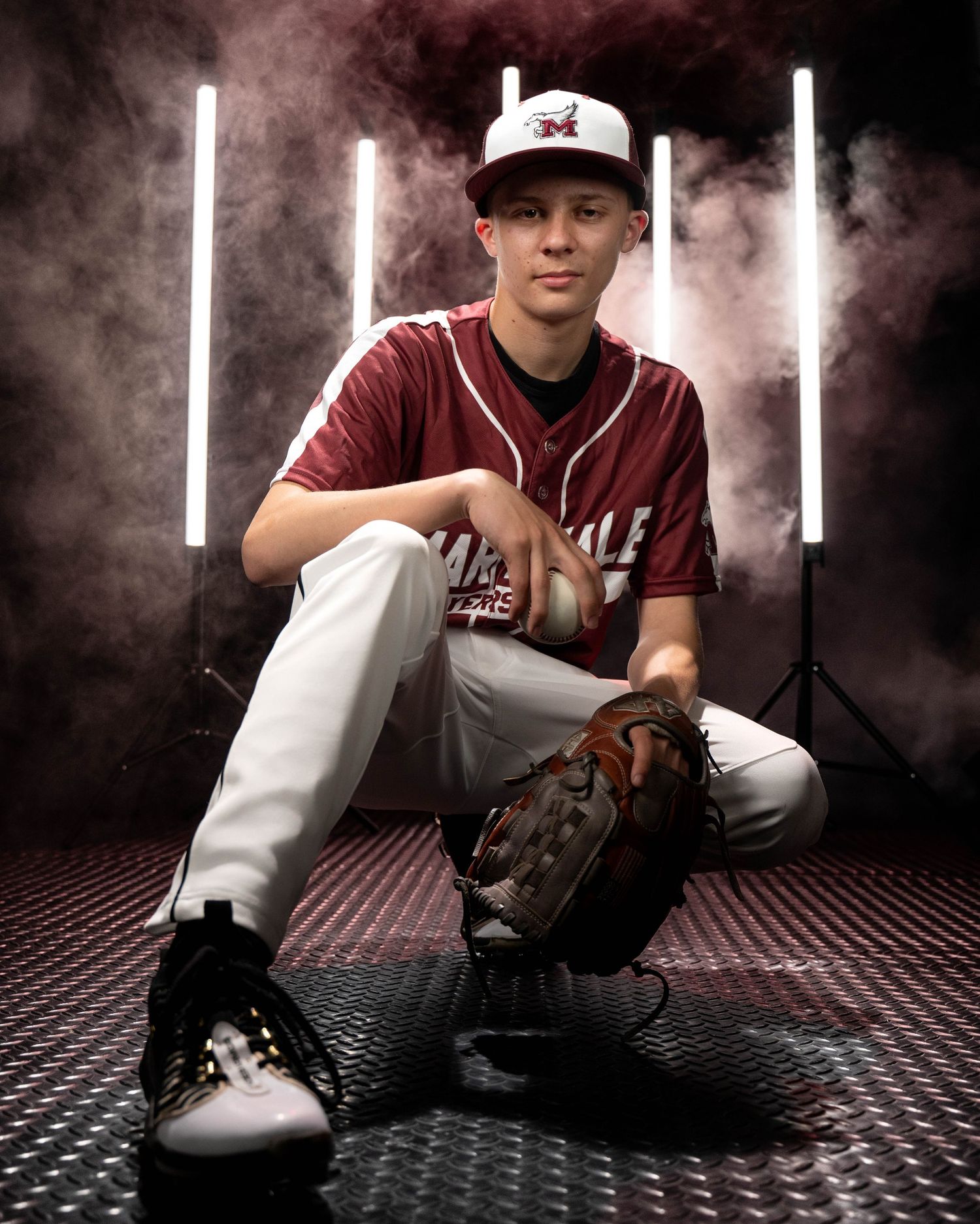 Teen baseball player in maroon uniform crouches with glove, posed against dramatic smoky studio backdrop with vertical lights.