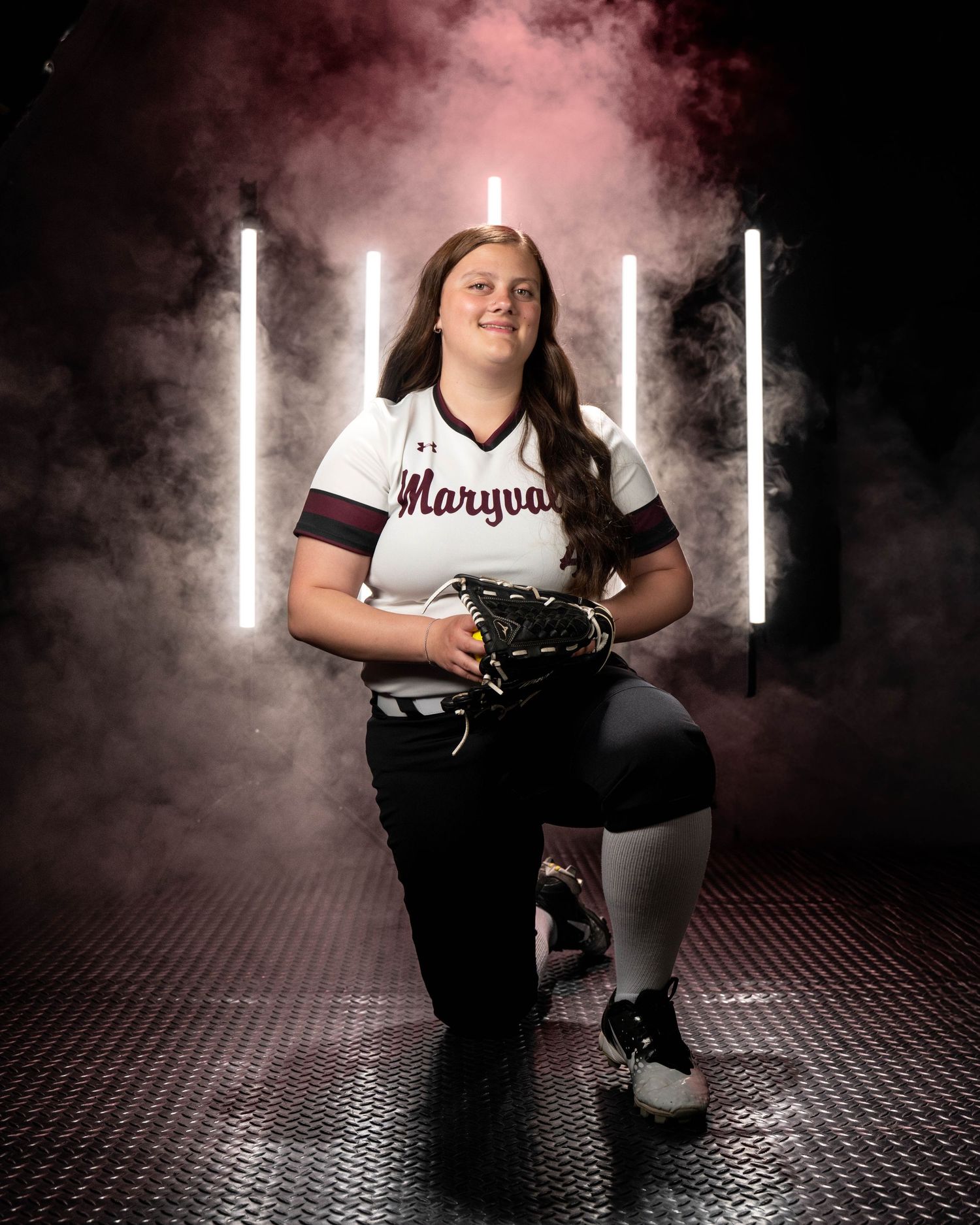 Female softball player in Maryvale uniform kneeling with glove in dramatic neon light and smoke setting.