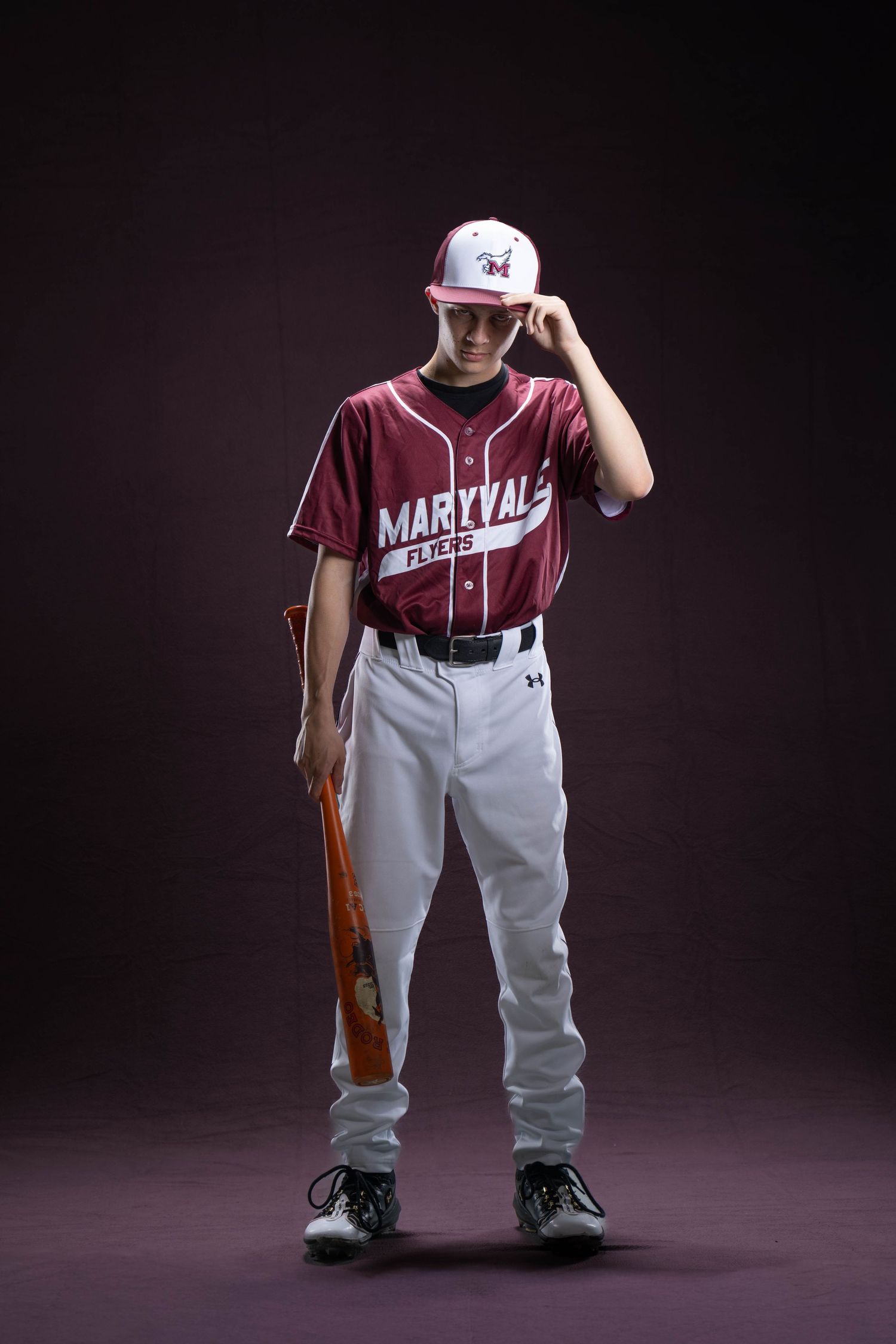 Young baseball player in Maryvale Flying uniform holding bat, wearing cap and cleats against dark background.