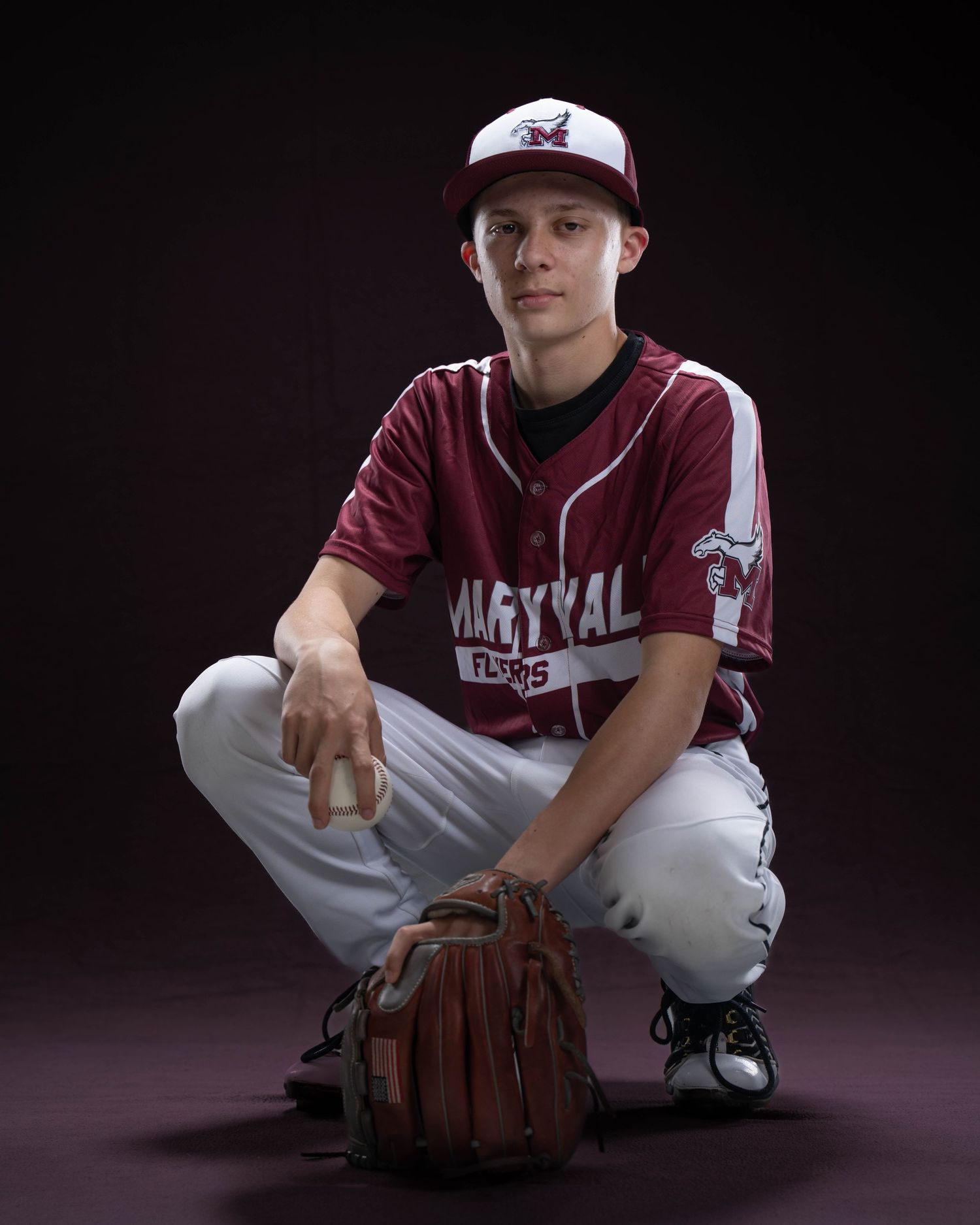 Young baseball player in maroon Maryval Flores uniform crouching with glove in dramatic studio portrait.