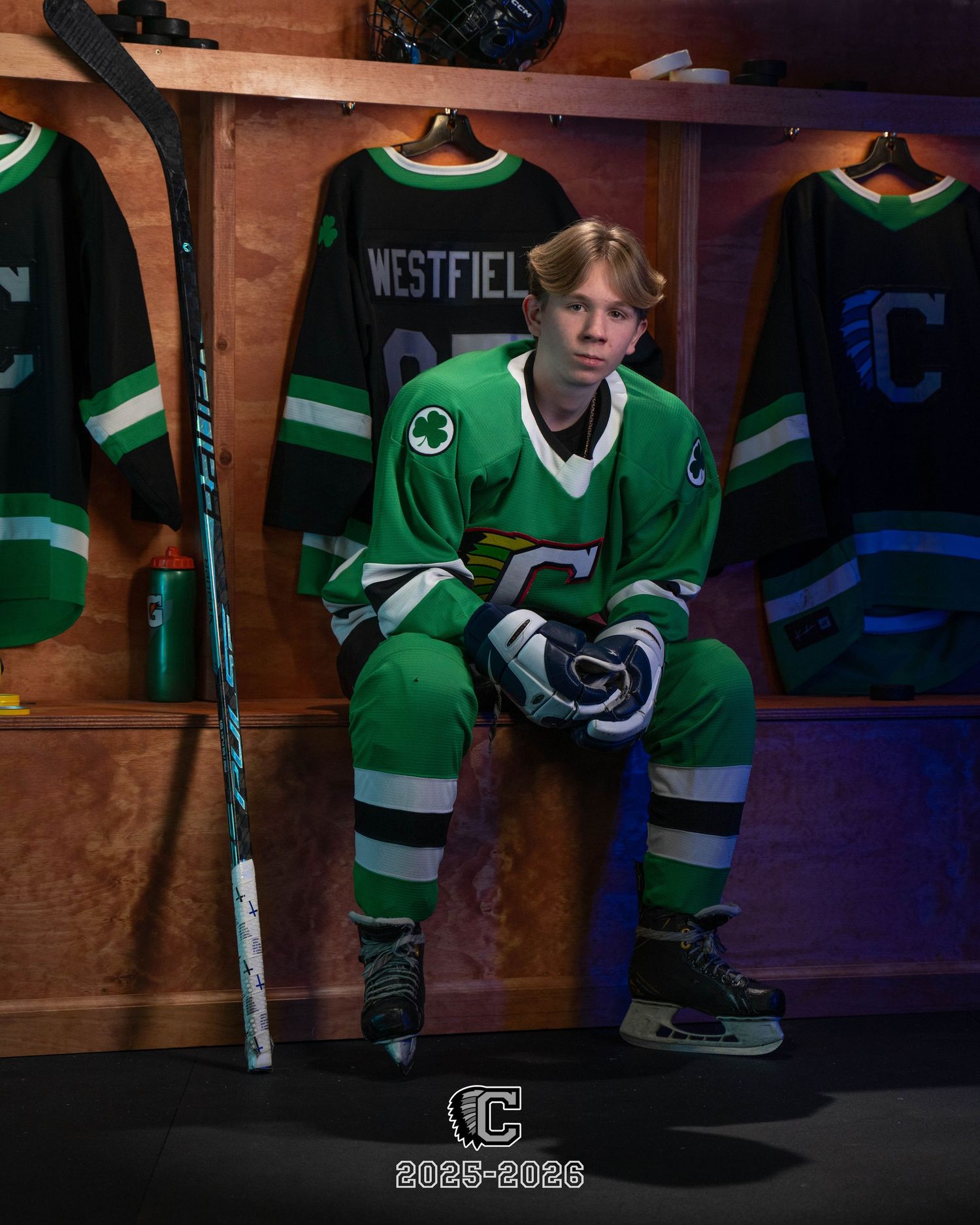 Hockey player in green jersey sits in locker room stall with equipment and gear visible in background.