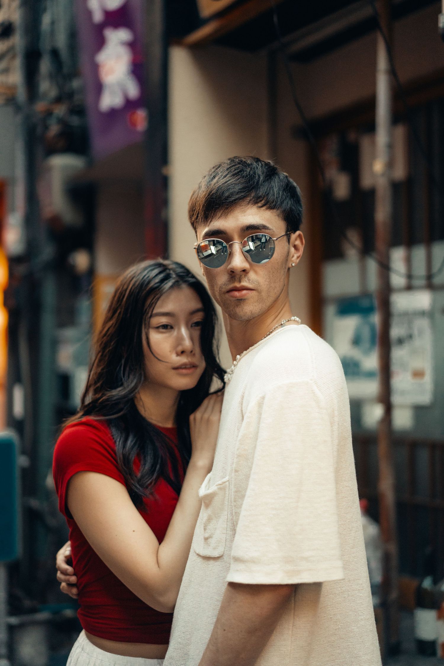 Stylish couple in white and red outfits pose together in an urban setting with atmospheric lighting and storefronts.