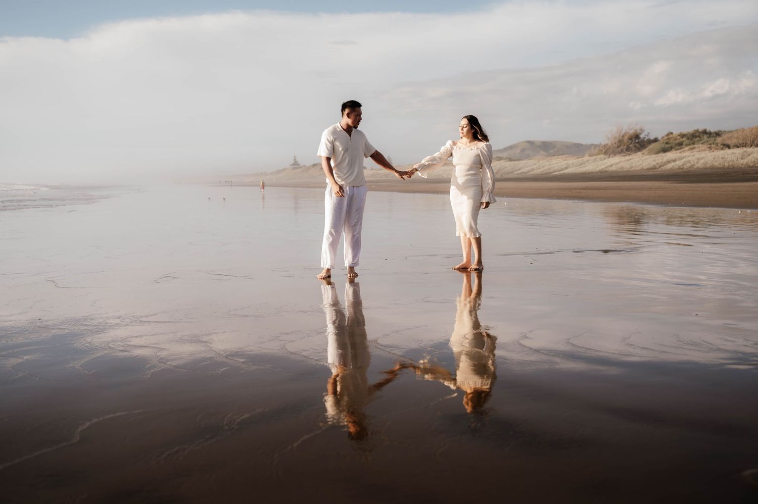Two figures hold hands while walking along a misty beach at sunset, creating reflections in the wet sand.
