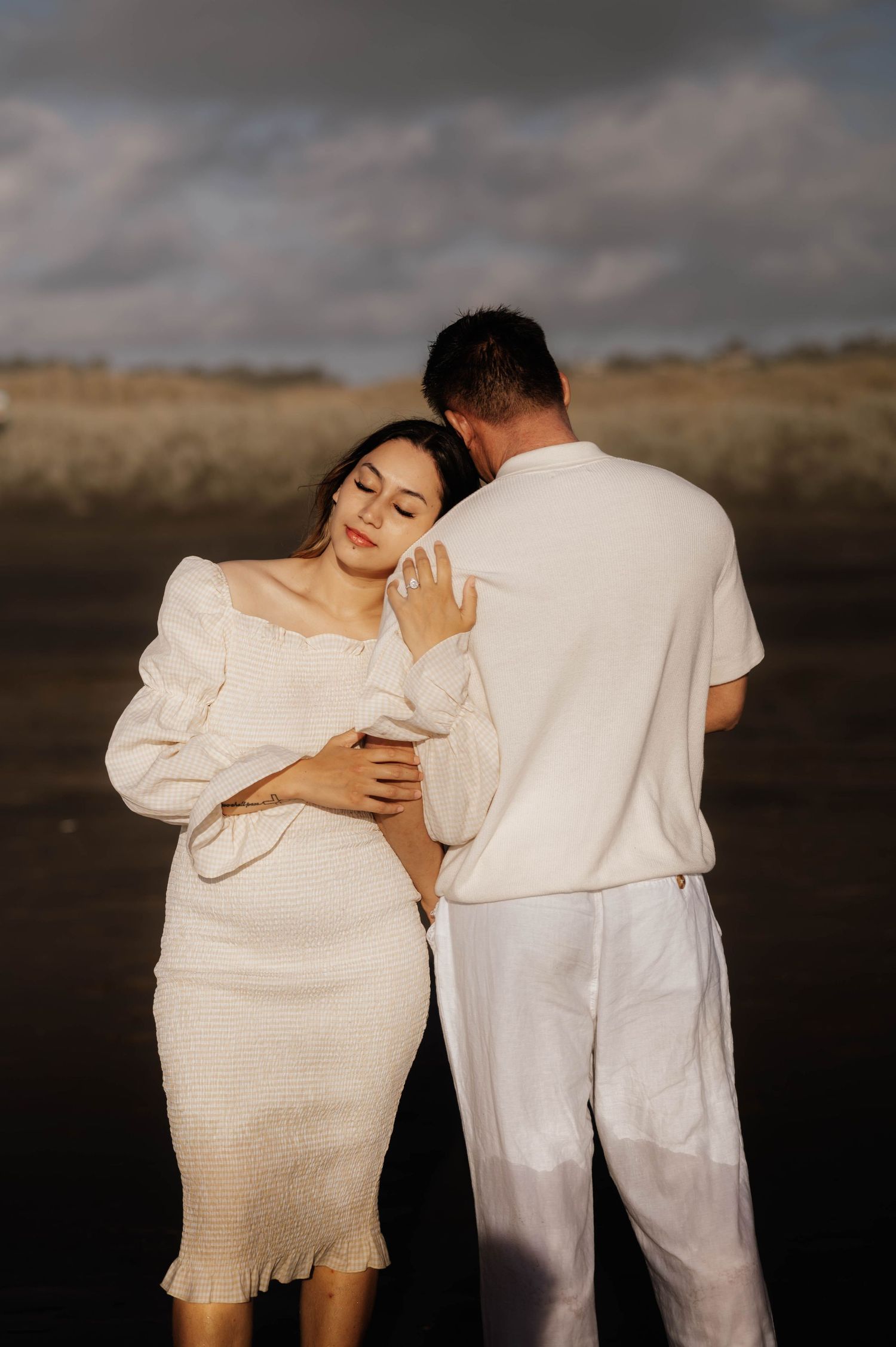 A romantic couple in white outfits embracing outdoors against a moody cloudy landscape.