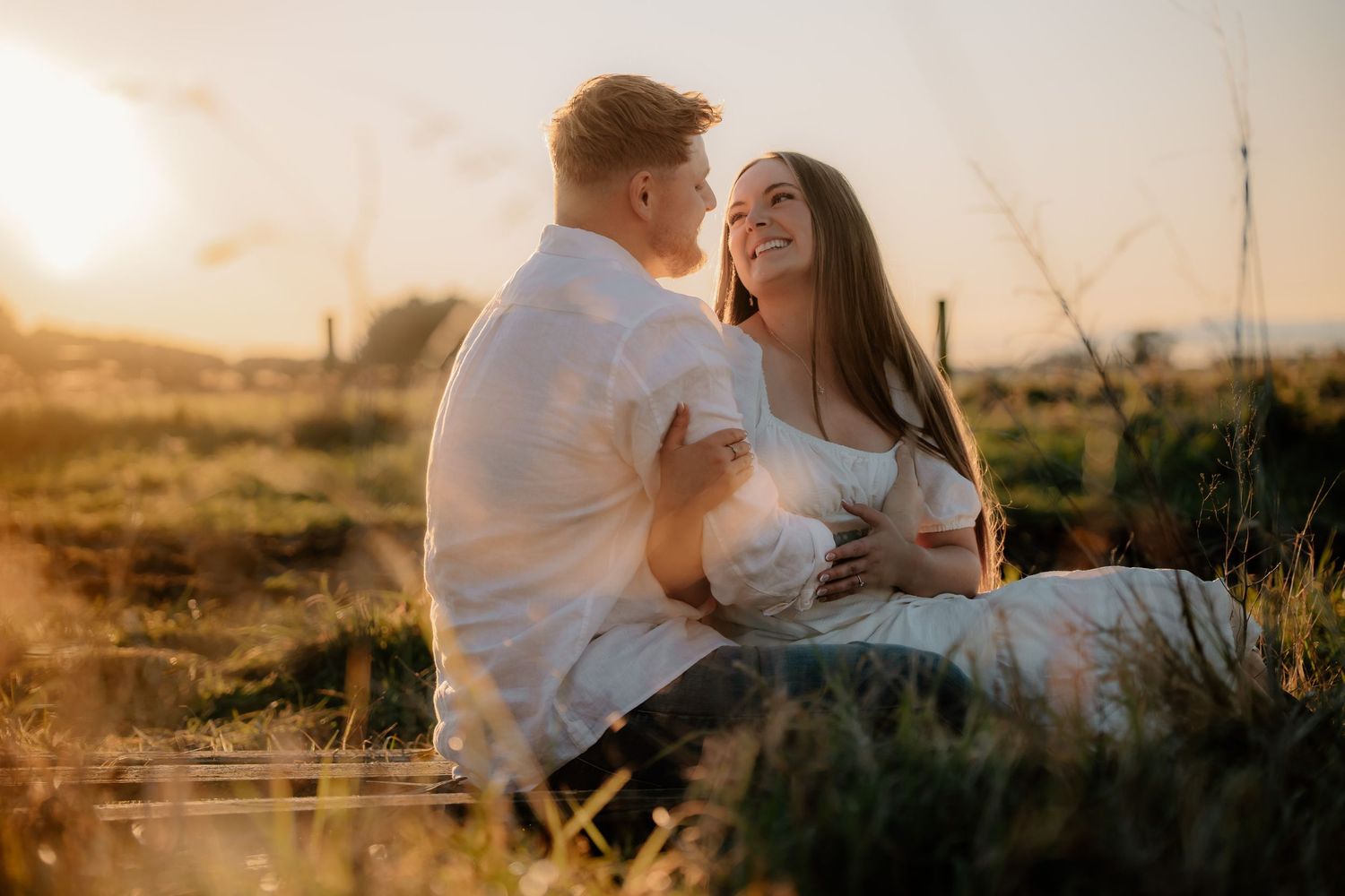 A romantic couple sits together in a sun-drenched field at golden hour, sharing an intimate moment.