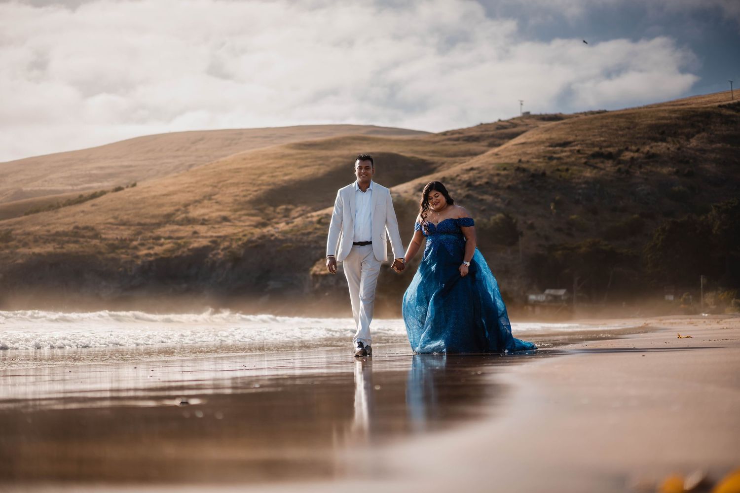 Two people in formal attire walk hand in hand along a misty beach with rolling hills in the background at sunset.