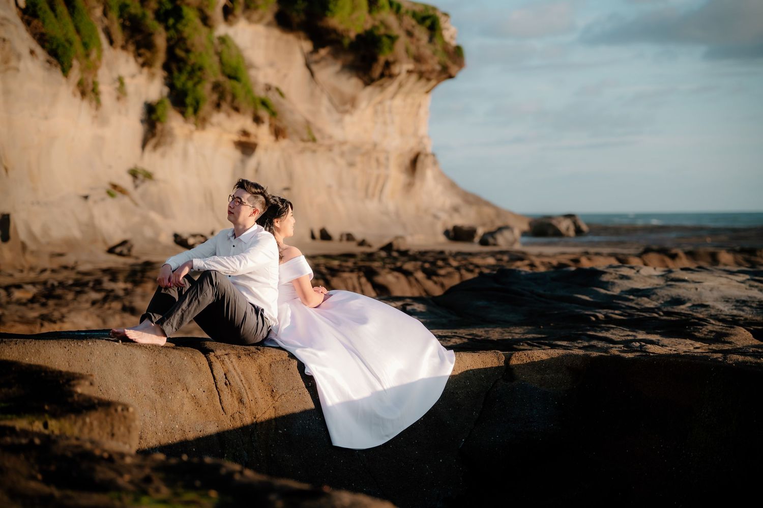 Two people in white shirts pose on a sandstone cliff overlooking an ocean view during sunset.