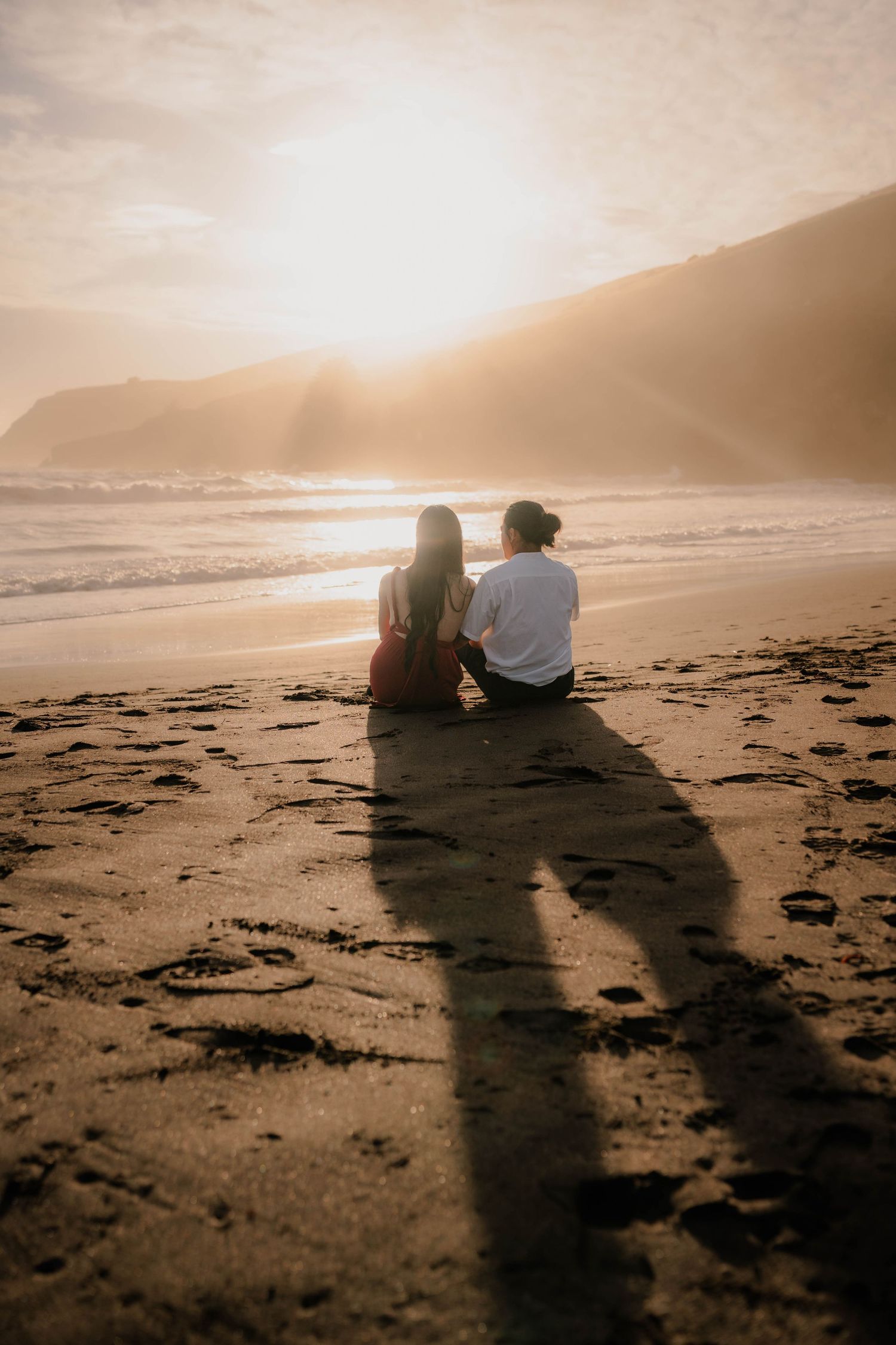 Two people sit together on a beach log at sunset creating long shadows on the sand while ocean waves roll in.