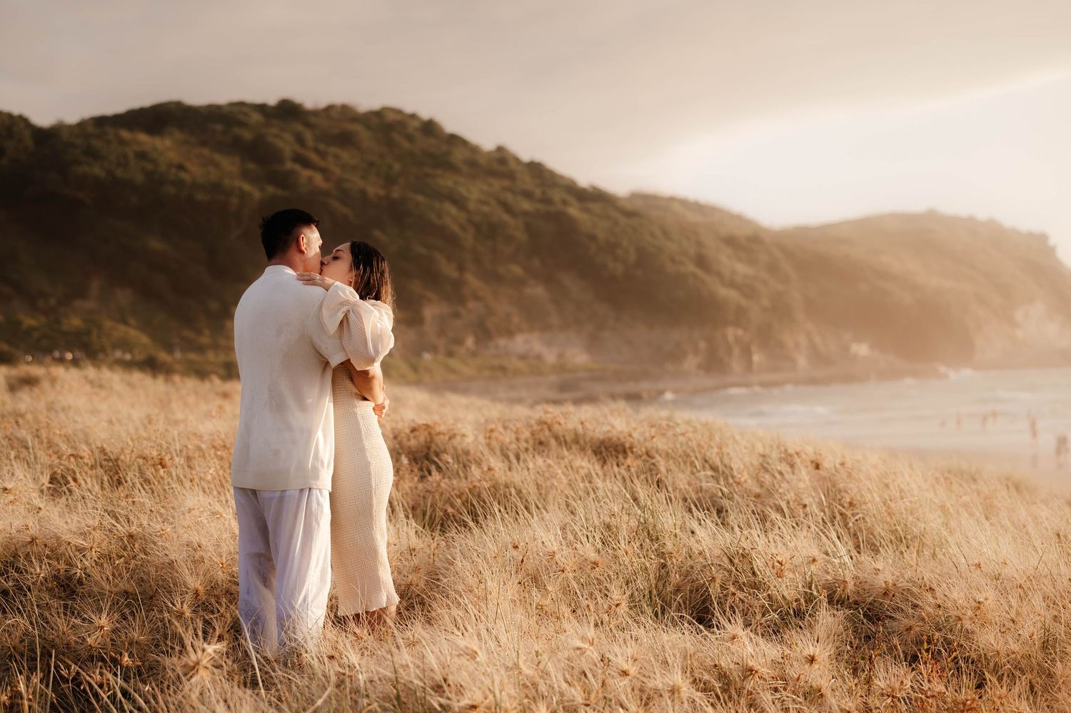 Romantic couple embrace at sunset on scenic coastal hillside with golden light streaming through tall grass meadow.