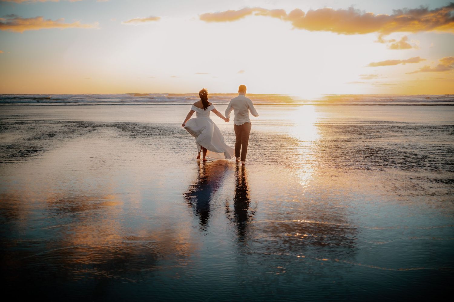 A couple walks along a beach at sunset holding hands while trailing a white wedding dress in the sand.