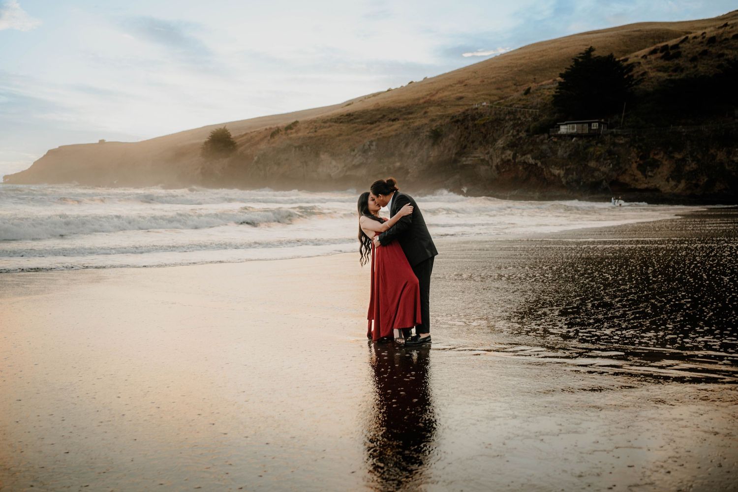 A person wearing a flowing red dress stands at the edge of a dramatic beach shoreline during golden hour.