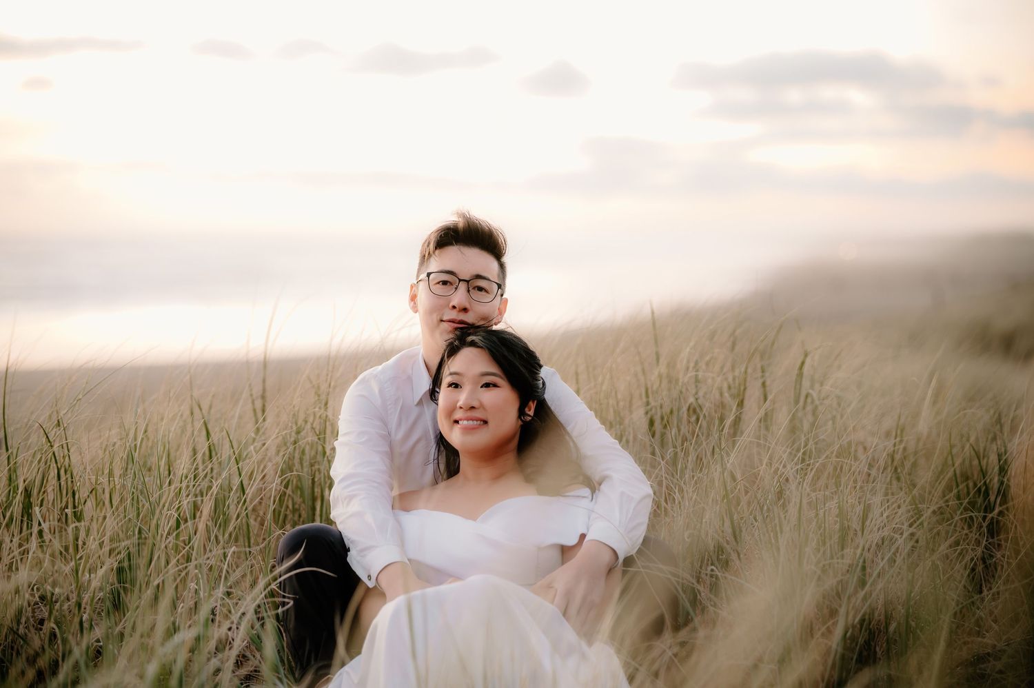 A couple in wedding attire stands in a golden field at sunset as their veil flows in the wind.
