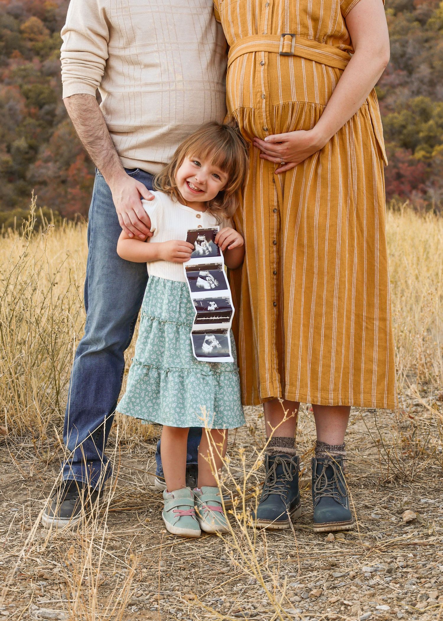 family photos in the utah fall mountains showing a mom hugging her pregnant belly and their daughter holding the ultrasound.