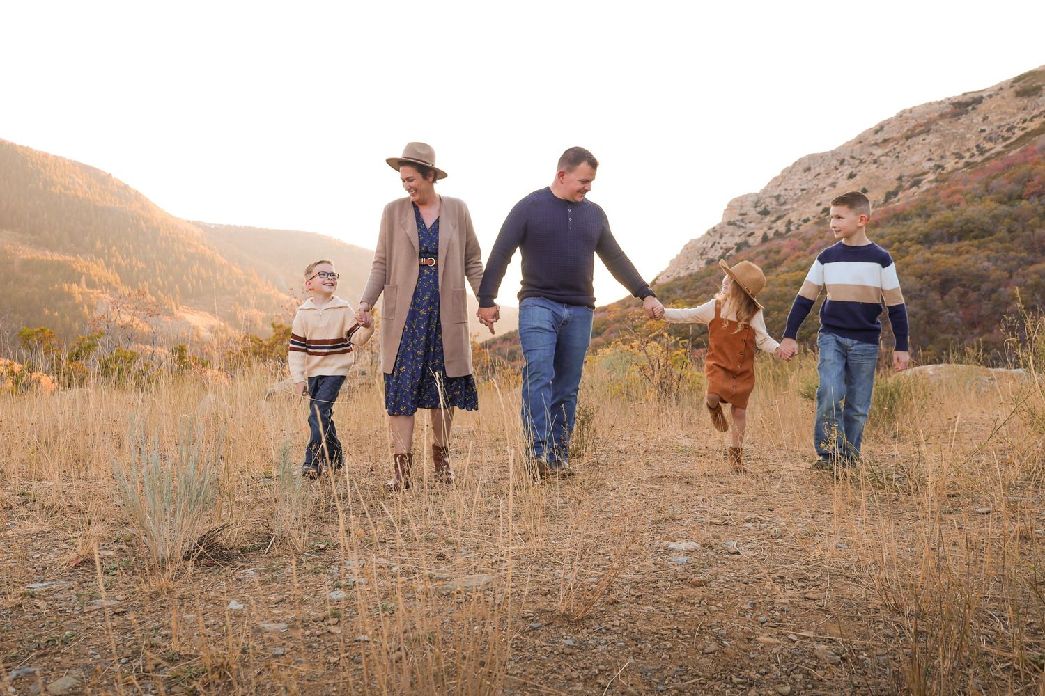 Family walking hand in hand outdoors during a fall photo session in the mountains, wearing cozy autumn outfits with scenic landscape