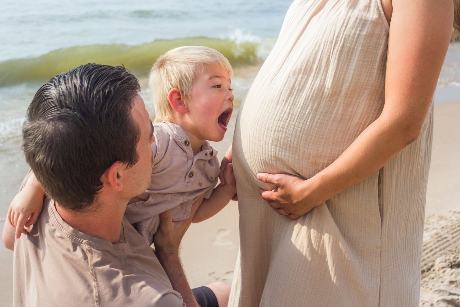 A maternity image of man holding his toddler son who is facing his mother's baby bump and attempting to give an open mouthed kiss.