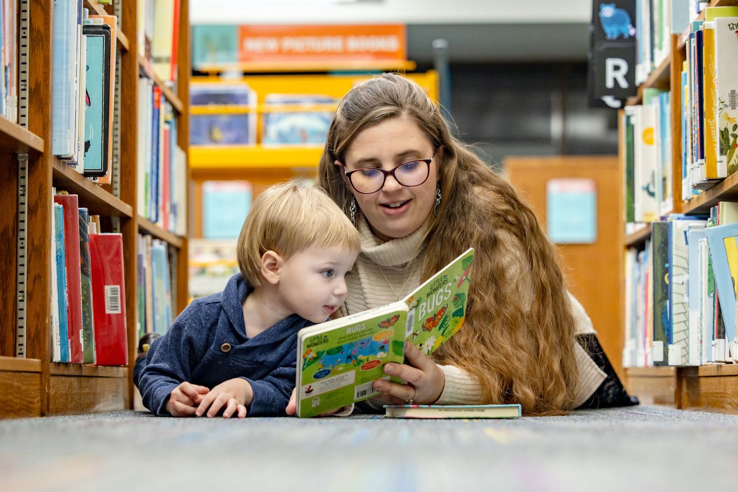 Picture of Mom and son laying on the floor between book shelves in a library looking at a book together.