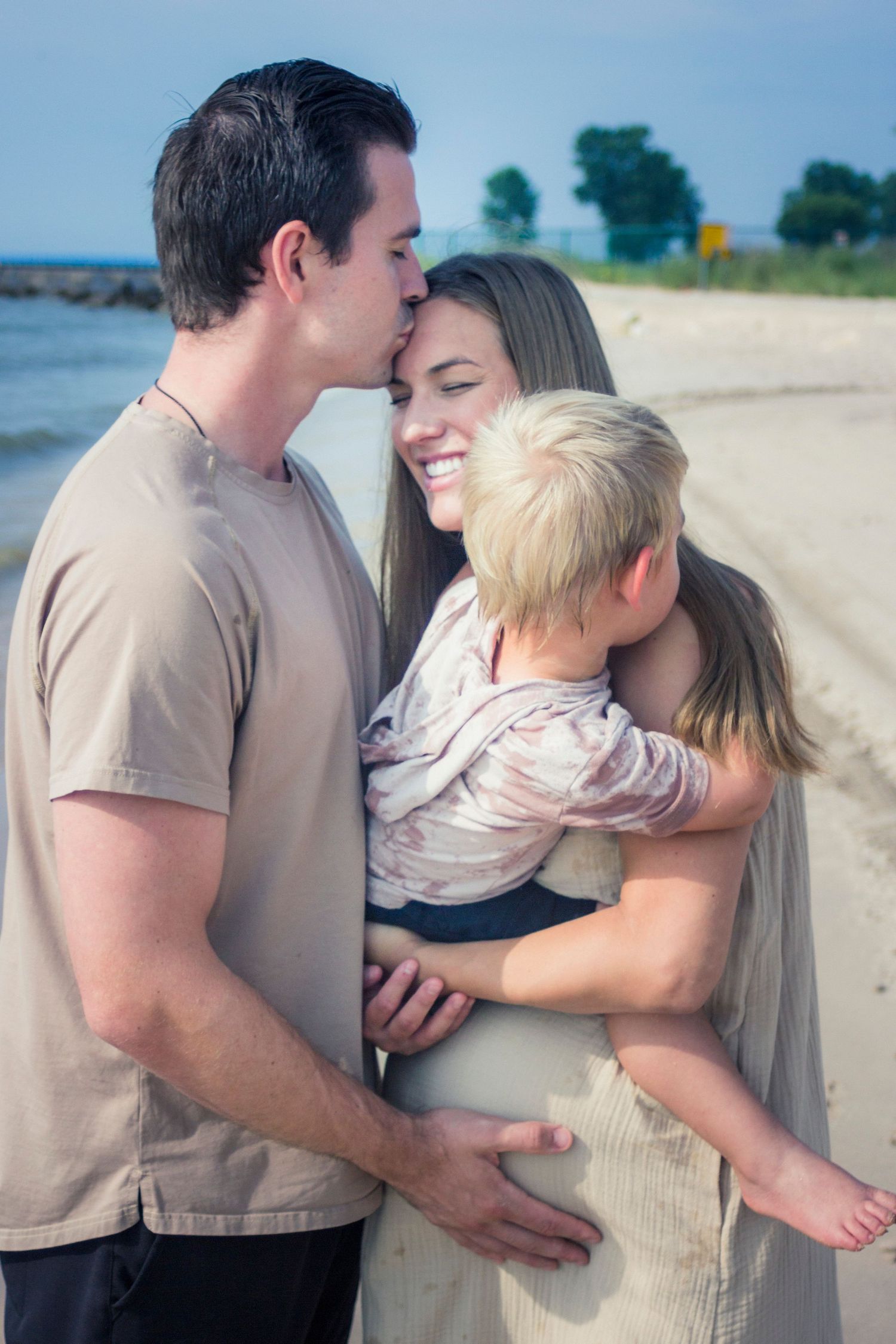 Maternity photos of a family on a beach with the husband kissing his wife who is holding their son.
