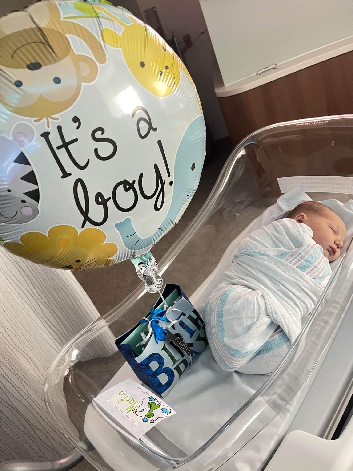 An image of a baby in a basinet in the hospital with a balloon that says 'It's a boy'.