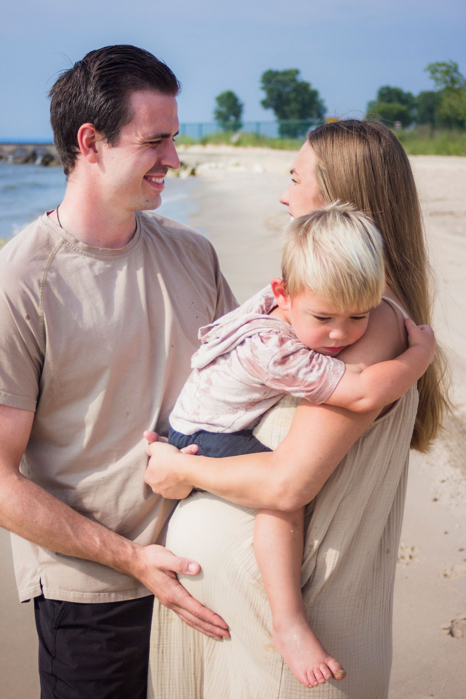 Maternity photos of a family on a beach with the husband looking at his wife and son.