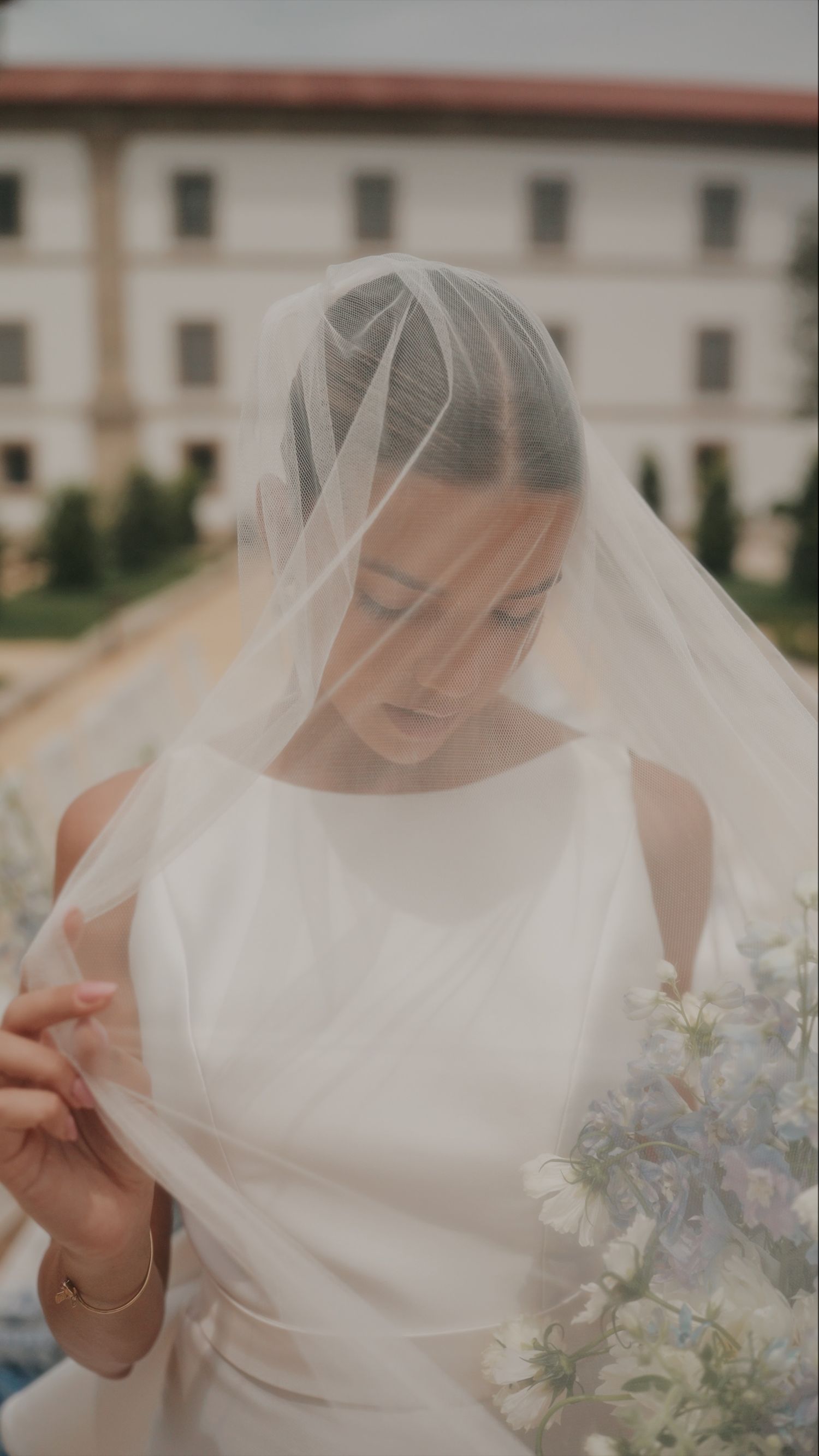 A bride in a classic white dress holds a delicate bouquet with soft blue flowers against a light background.