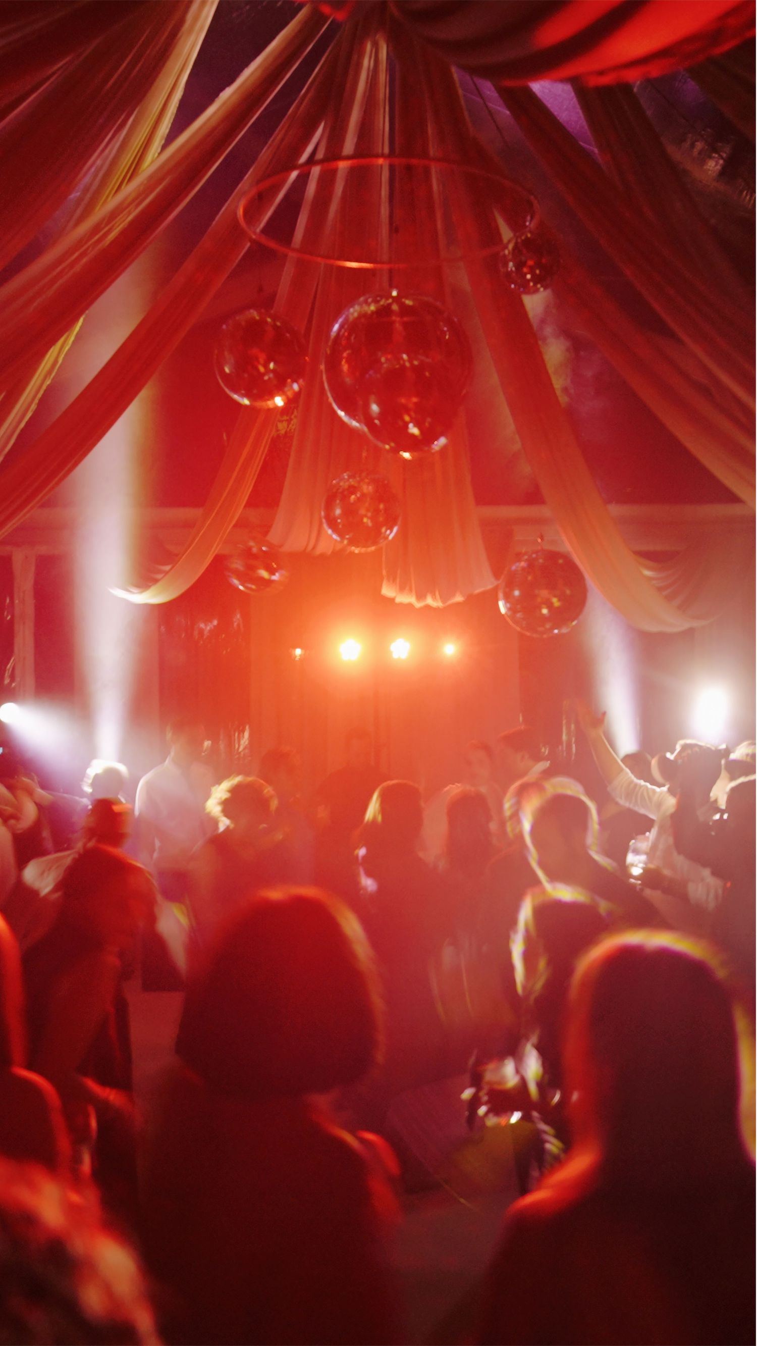 Red-lit party scene with hanging glass orbs and crowd silhouettes under draped fabric ceiling.
