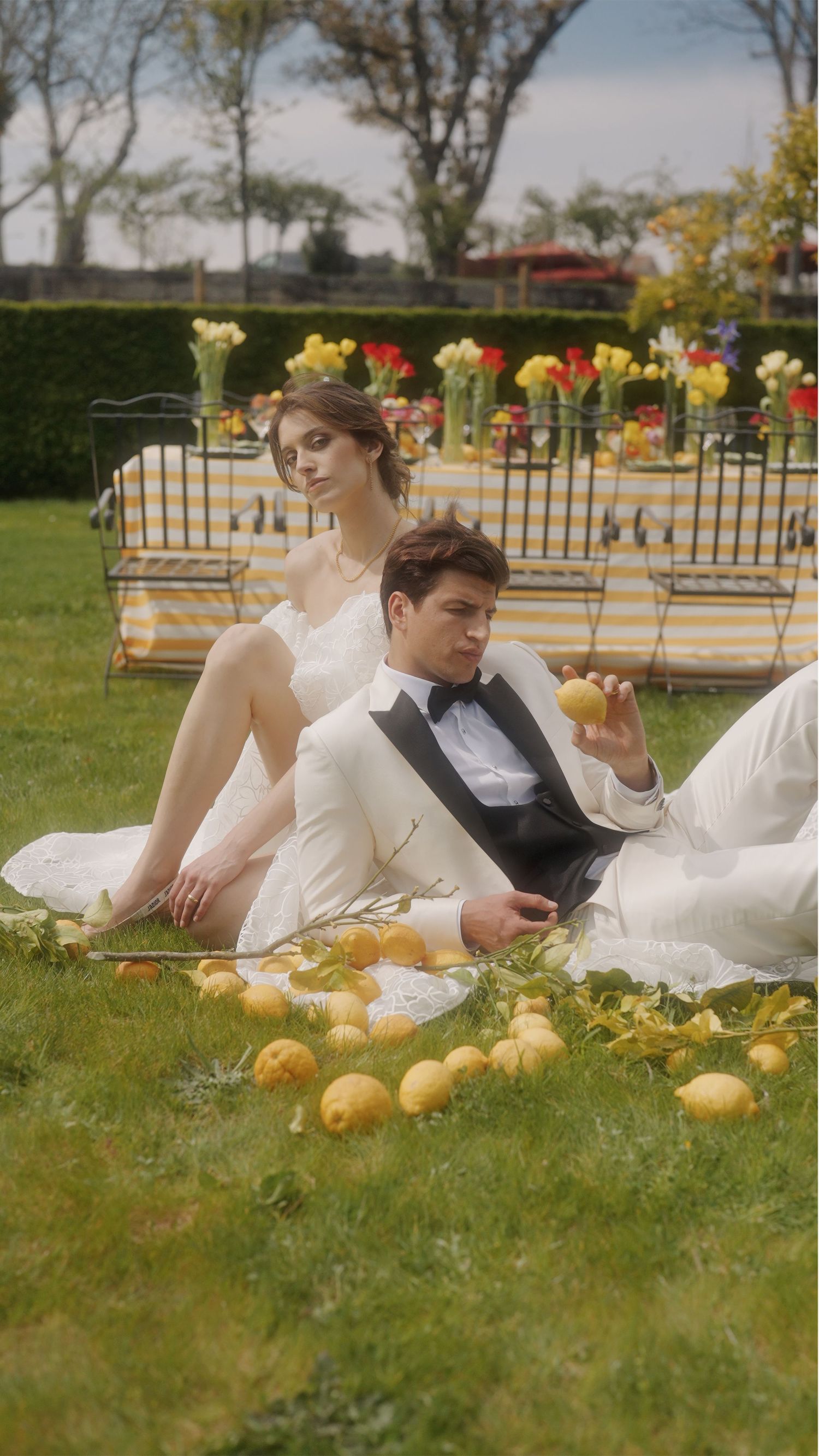 Romantic wedding scene with couple lounging on grass surrounded by lemons and flowers.