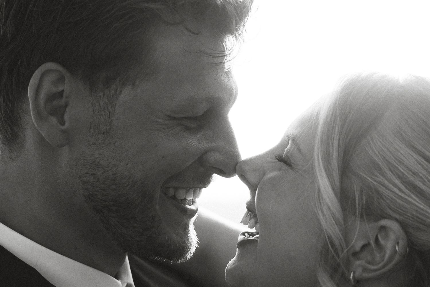 Black and white photo of a smiling couple sharing a romantic moment, nearly kissing, bathed in soft natural light.