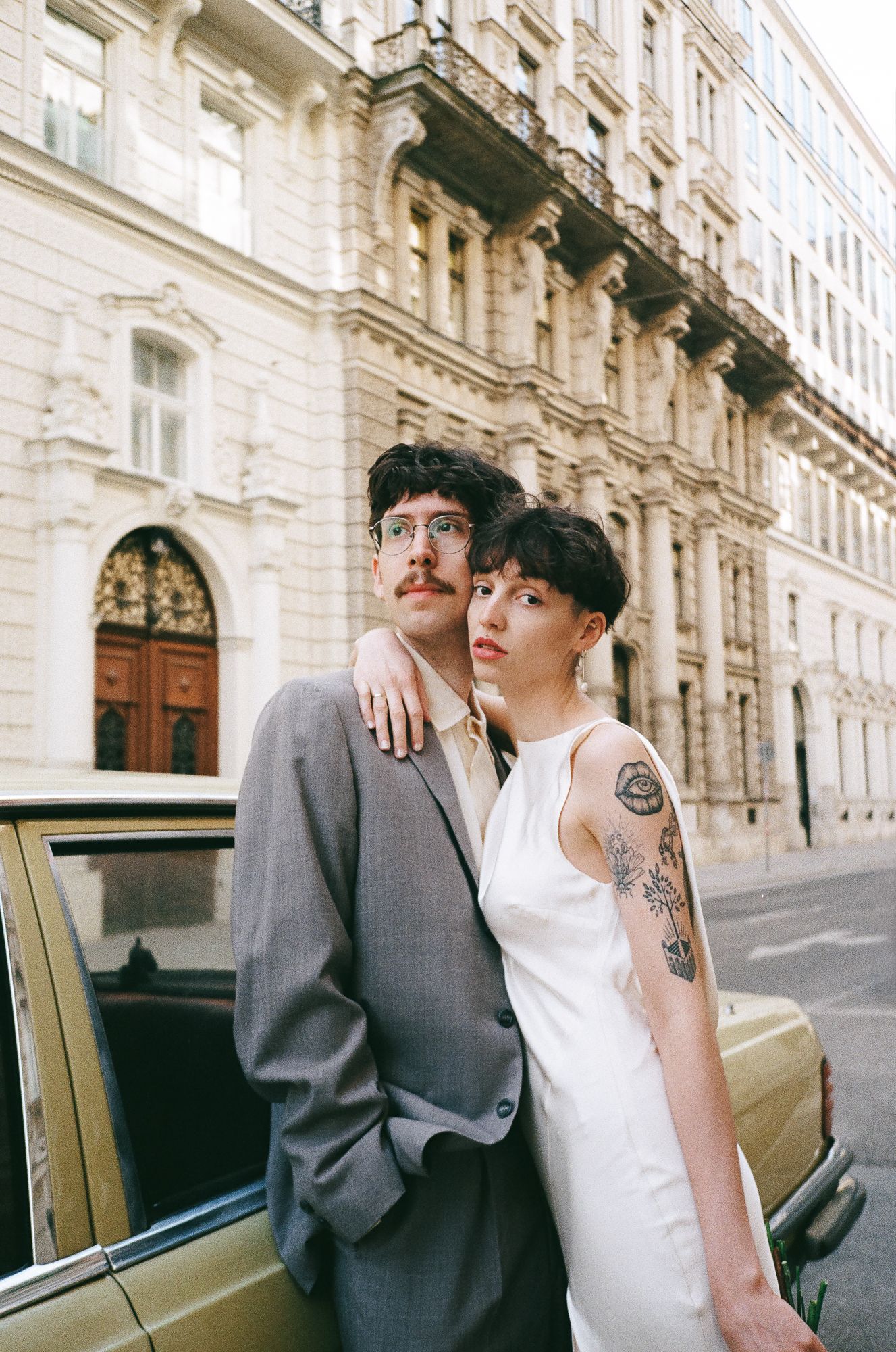 A couple in wedding attire poses by a vintage car on a European city street, the woman showing arm tattoos.