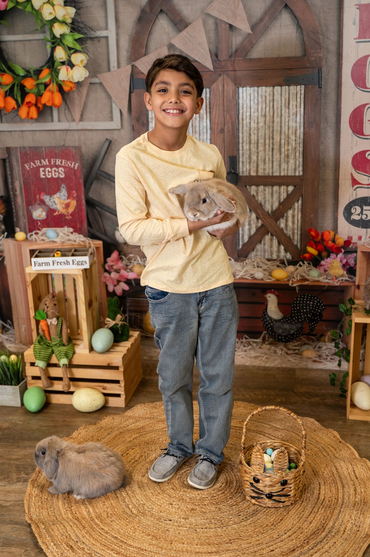 Child in yellow sweater and jeans poses with Easter decorations and bunny on round jute rug in rustic setting.